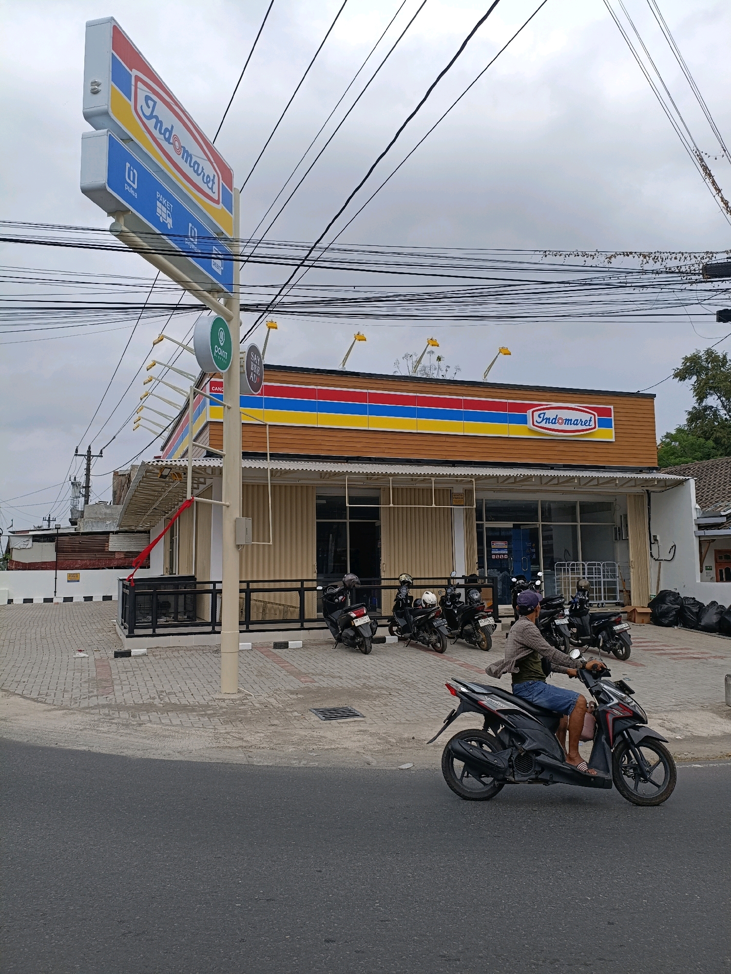 A low-angle, full shot captures the exterior of an Indomaret convenience store in Purwomartani, Indonesia, under an overcast sky. A prominent red, blue, and yellow Indomaret sign stands tall on the left, with additional signage below indicating services like "pulsa" (credit) and "paket" (package). The store itself has a brown, wood-textured facade with a white awning and large glass windows. Rows of parked motorcycles are visible in front of the store, and a person wearing a plaid shirt, blue shorts, and sandals rides a black motorcycle towards the foreground, moving from right to left across the frame. Black trash bags are piled up to the right of the store entrance. The ground is paved with interlocking bricks and concrete. Overhead, a dense network of electrical wires crisscrosses the sky.