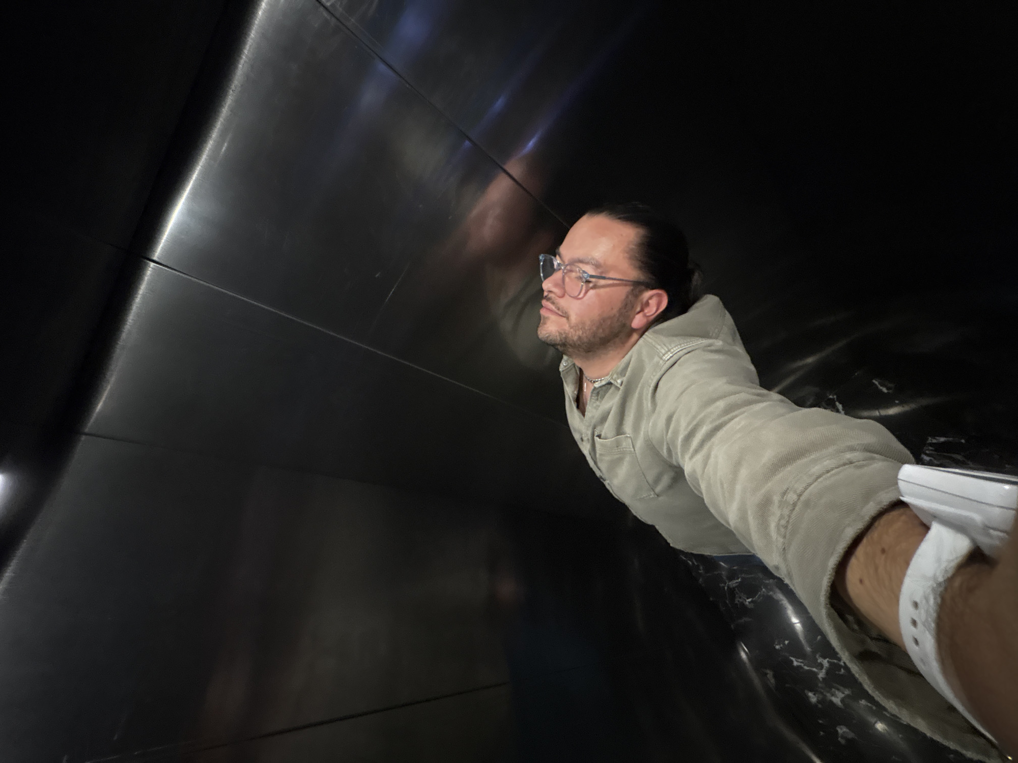 A man wearing glasses and a light-colored button-down shirt is inside a dark, enclosed space lined with black metal panels. The man has his hair pulled back and a wristband on his left wrist, suggesting a recent hospital visit. The environment appears to be a modern architectural structure, possibly an art installation or a building interior. Based on the city mentioned, Puebla, Mexico, the location could be the International Museum of the Baroque.