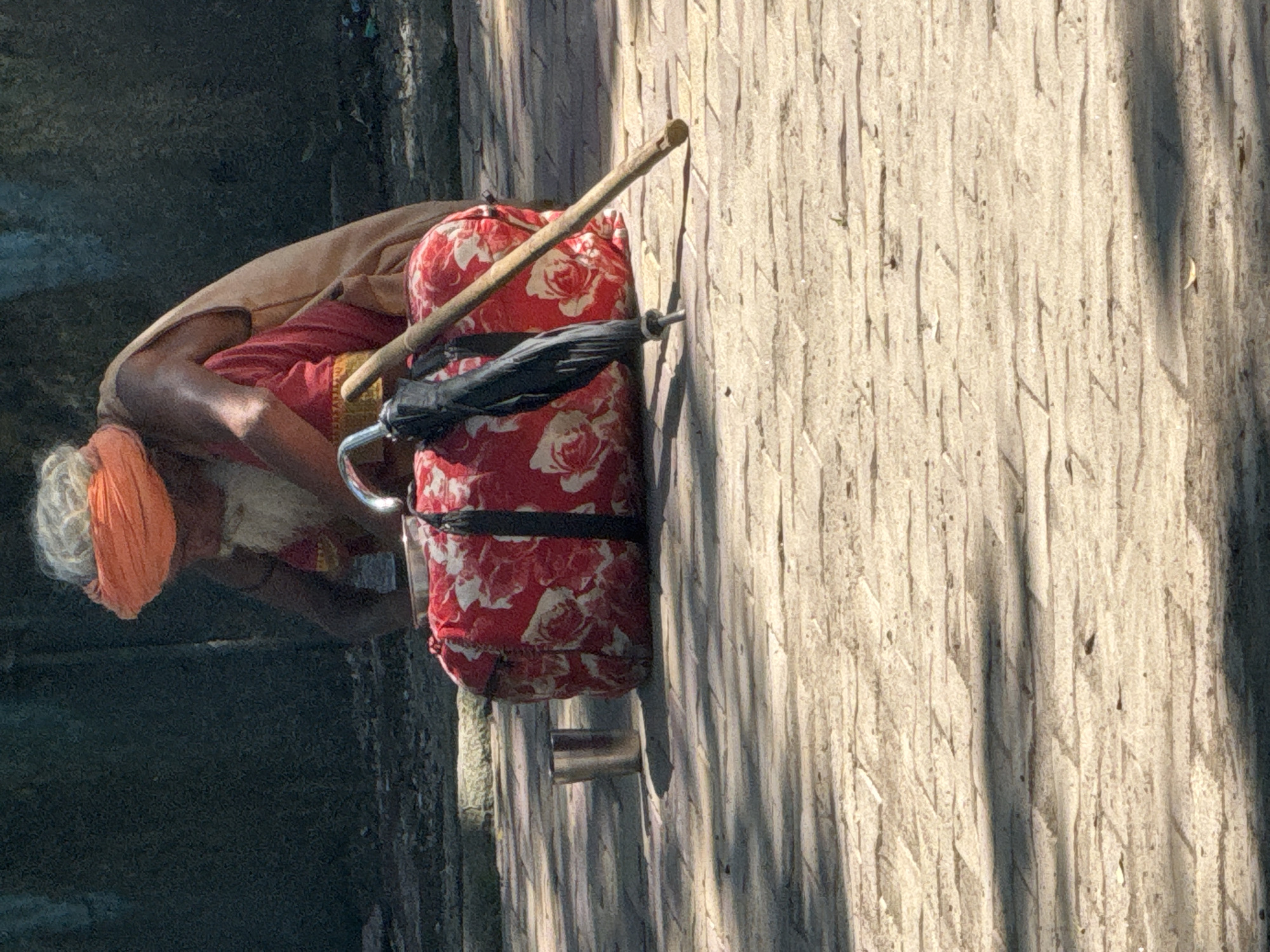 In Rishikesh, India, a man with grey hair and a white beard wears an orange turban and a red garment. He is sitting with a large duffel bag with a red floral pattern in front of him. An umbrella and a wooden stick lean against the bag. A metallic cup sits to the left of the bag. The ground is paved with stone tiles, and shadows from surrounding trees are cast across the surface. The background shows a dark, textured wall.