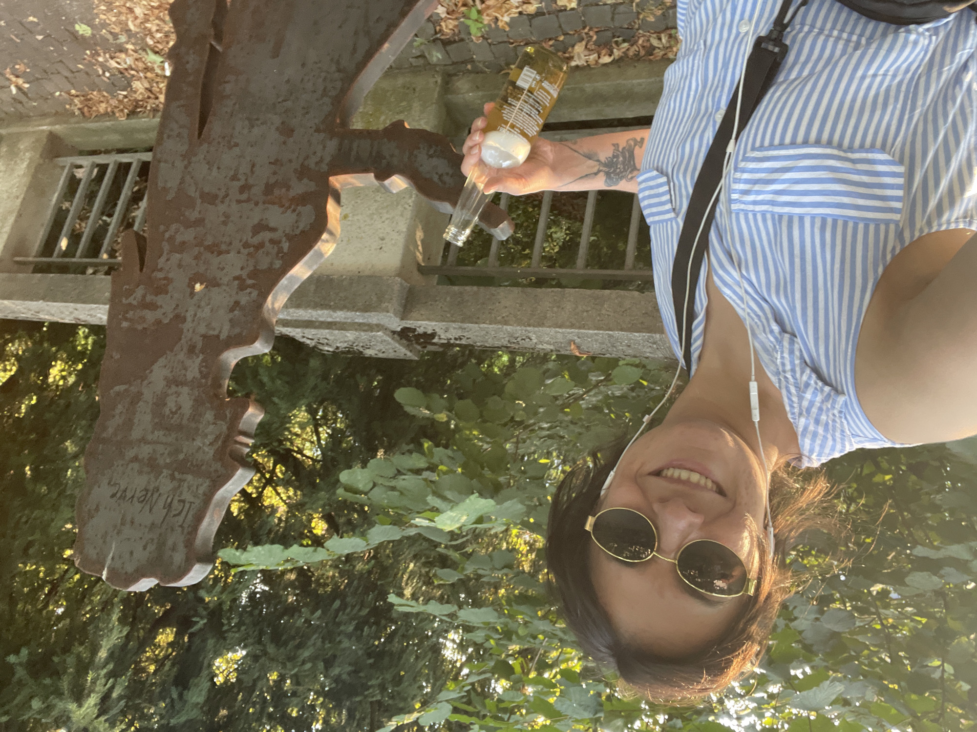 A woman with dark hair, wearing sunglasses and a blue and white striped shirt, smiles at the camera and holds a beer bottle. She is standing next to a metal sculpture of a human head. The sculpture has the words "Ich Nerve" inscribed on it. The background shows trees and foliage, suggesting an outdoor setting. The scene appears to be in Iserlohn, Germany, near the sculpture "Ich Nerve" by the artist Horst-J.H. Piller.