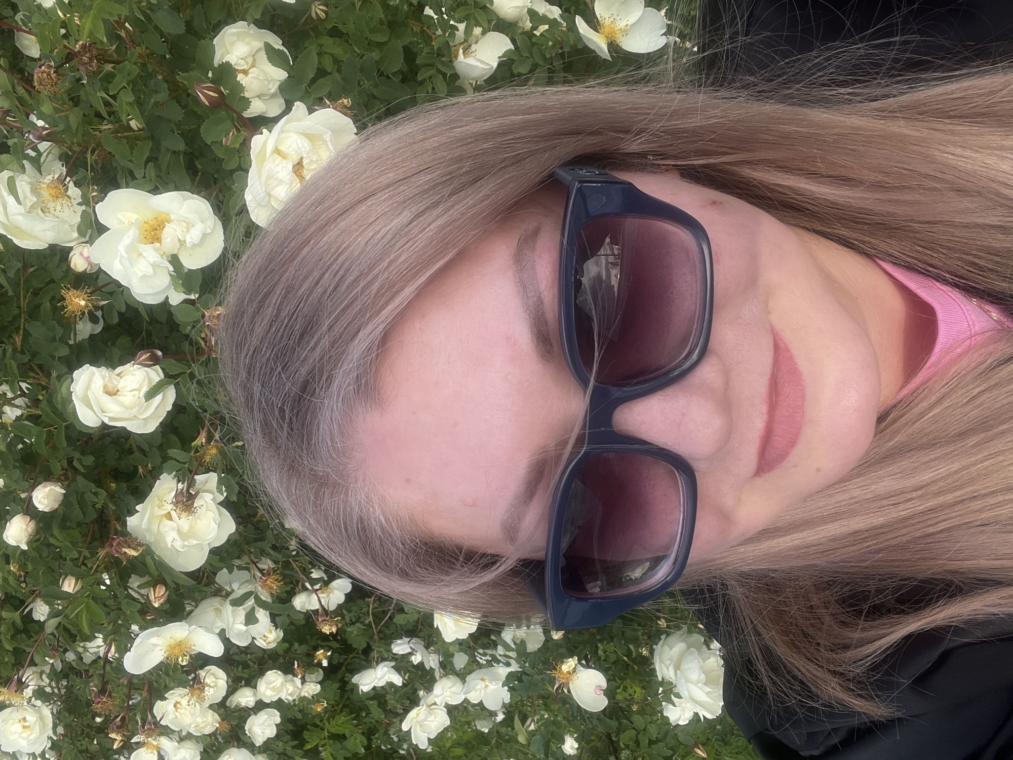 An angled, close-up selfie captures a person with long, light blonde hair, streaked with grey or silver near the roots, partially obscuring their face. The person is wearing dark-framed sunglasses with reflective lenses, beneath which a hint of pink lipstick is visible. A pink garment can be seen near the neck, with a black outer layer visible on the right. The background is a dense bush filled with numerous white, open-faced flowers, likely roses, characterized by their delicate petals and prominent yellow stamens, interspersed with green leaves. Some withered flowers and buds are also present. The scene is brightly lit by natural daylight, suggesting a sunny or overcast day. The composition implies a relaxed pose, possibly lying down or tilting the head to capture the surrounding flora. No visible text is present. The context provided places this scene in Shud'ya, Russia.
