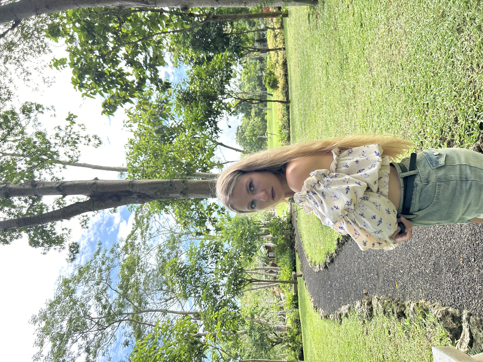 An image, oriented vertically as if taken in portrait mode and then rotated 90 degrees clockwise, captures a young woman standing in a lush outdoor setting. The woman, with long, straight blonde hair parted down the middle, looks directly at the camera with a neutral expression. She is wearing a light-colored, off-the-shoulder crop top featuring a delicate blue and white floral pattern with ruffled sleeves. Her attire also includes light-wash denim shorts cinched with a dark belt, which her right hand is touching.
The background presents a vibrant natural environment, indicative of a park or garden. Expansive areas of well-maintained green grass dominate the scene, interspersed with numerous tall trees boasting dense green foliage. A dark, textured path, appearing to be made of small stones or gravel, winds through the grass on the right side of the frame, leading further into the distance. Above, through the tree canopy, patches of bright blue sky with scattered white clouds are visible, suggesting a clear, sunny day. The overall impression is serene and natural. No specific activity is depicted other than posing for the photograph. There is no visible text in the image. The provided location context is Kuta Selatan, Indonesia.