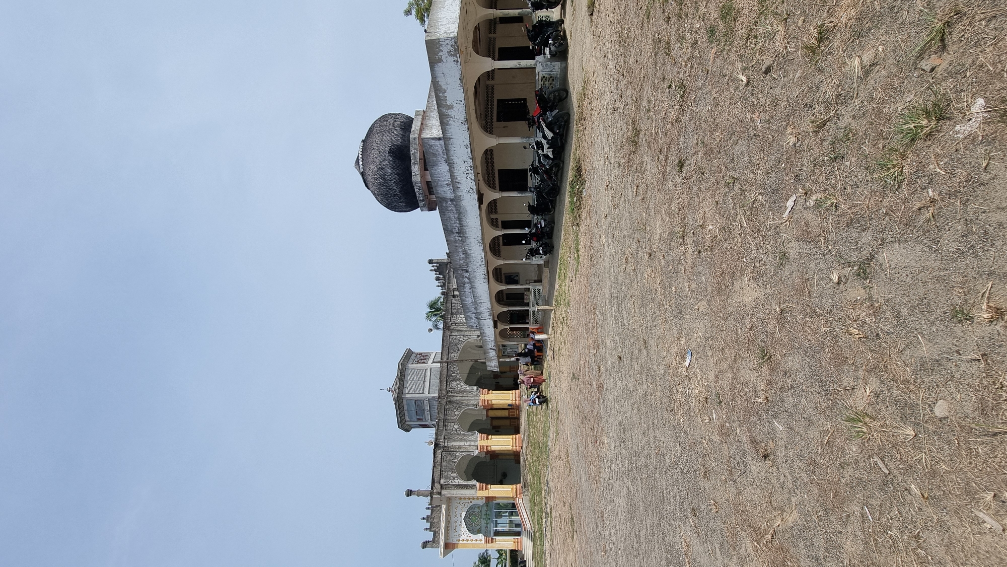 The image shows the exterior of a building, likely a mosque, with a courtyard in front. The building features multiple arches and a dome. Several motorcycles are parked in front of the building. A few people are gathered in the courtyard, some standing and others seated. The ground is covered with dirt and dry vegetation, suggesting a warm, dry climate. The sky is clear and blue. The building appears to be located in Tumpokbeurandang, Indonesia, given the provided context.