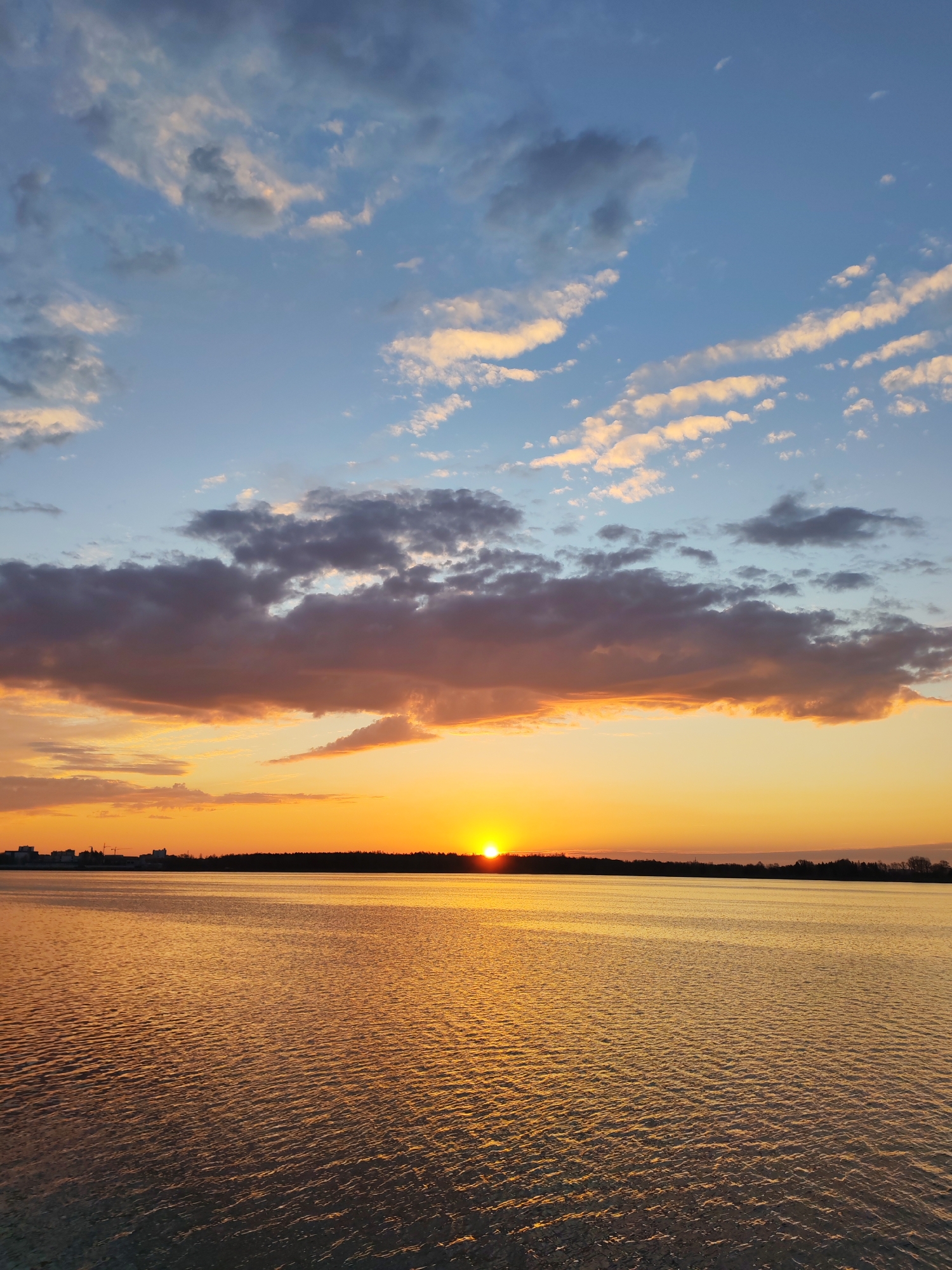 The image depicts a sunset over a body of water, likely a lake. The sun is setting on the horizon, casting a warm golden glow on the water. The sky features a mix of blue and orange hues, with scattered clouds, some tinged with pink and purple. A dark treeline is visible on the horizon, separating the water from the sky. On the left side of the treeline, there are buildings. The water is calm with slight ripples. The image was taken in the city of Unknown, Unknown.