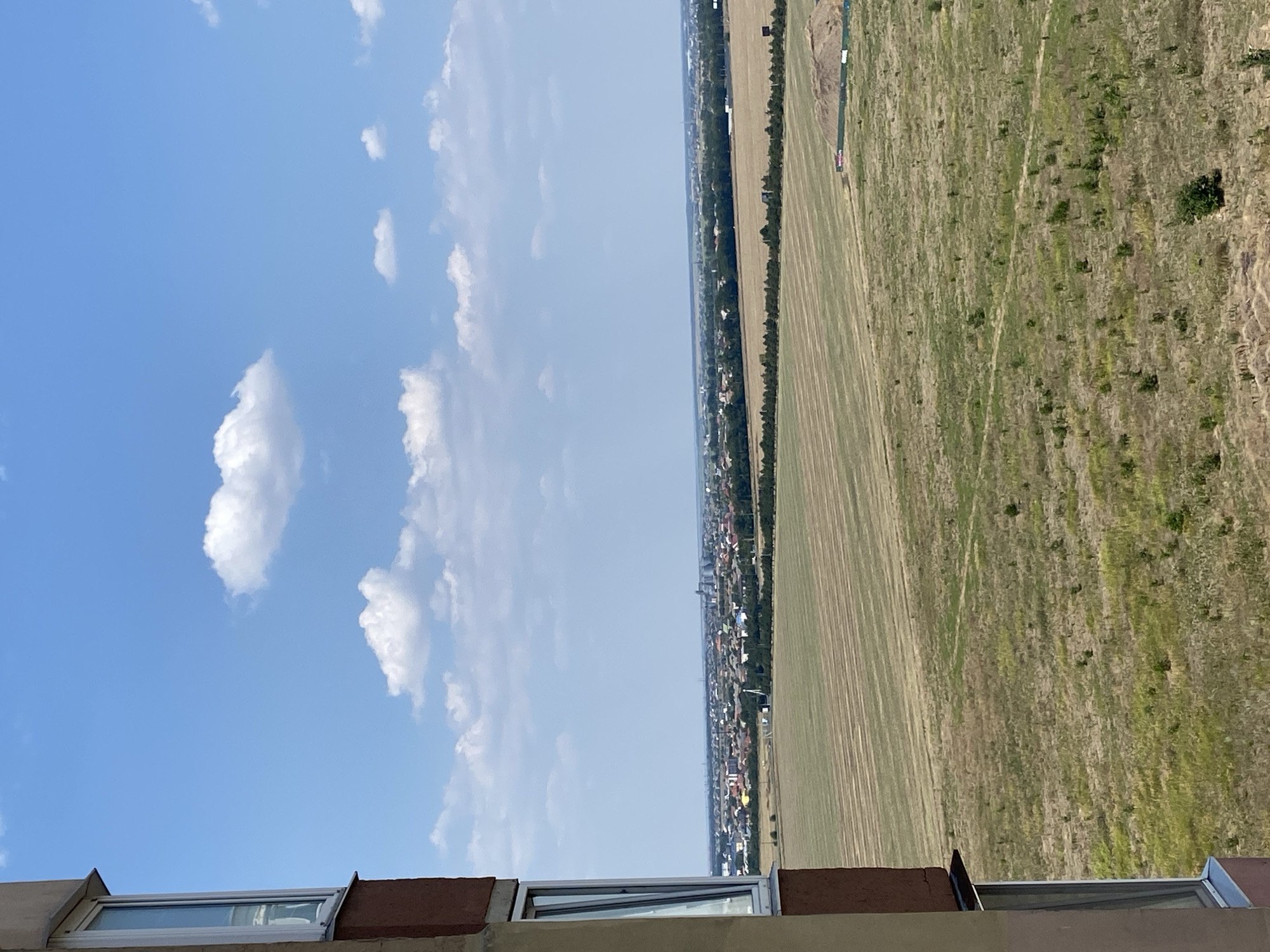 The image depicts a vast, sun-drenched landscape viewed from a high vantage point, likely an apartment building. In the foreground, the corner of a building with a partially open window is visible on the left, its brown and beige facade contrasting with the bright sky. The midground reveals an expansive agricultural field, characterized by parallel lines suggesting recently harvested crops or tilled land. This field transitions into a more natural, grassy area dotted with small shrubs and trees.
Beyond the fields, a sprawling urban area stretches across the horizon. A dense collection of buildings, appearing to be residential homes and possibly some industrial structures, forms a distinct townscape. A line of trees acts as a natural boundary between the fields and the settlement. In the distance, the sky is a clear, vibrant blue, with a few scattered white cumulus clouds adding texture to the scene. The overall impression is one of a sunny day in a rural or semi-rural setting, possibly on the outskirts of a city. The provided location information suggests this scene is situated in Chornomorsk, Ukraine.