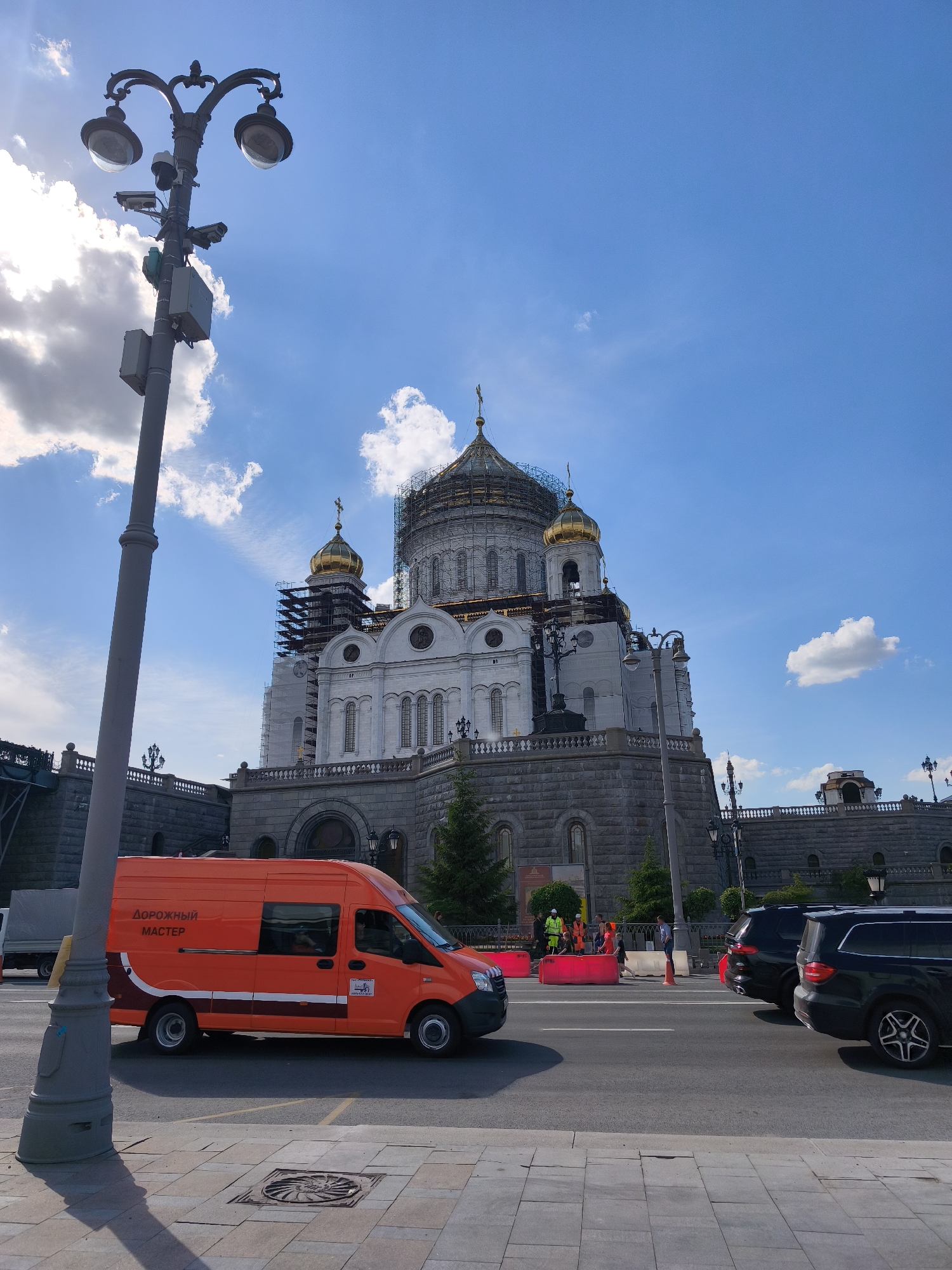 An outdoor, eye-level shot captures a vibrant urban scene dominated by a grand white church with golden domes, partially covered in scaffolding, indicating ongoing construction or renovation. The church, possibly a cathedral, has a large stone base and intricate architectural details.
In the foreground, a prominent black lamppost with an ornate, branching design stands on the left, equipped with multiple surveillance cameras and other electronic devices.
An orange commercial van is parked on the street in the middle ground, facing left. The side of the van clearly displays the text "ДОРОЖНЫЙ МАСТЕР" (Dorozhny Master, meaning "Road Master" or "Road Worker" in Russian). Behind the van, two dark-colored SUVs are visible, parked on the right side of the street.
Further back, near the base of the church, several construction workers or public service personnel in high-visibility yellow and orange clothing are gathered around red plastic barriers. A small tree is also visible near the church's base. The street has clear markings, and a manhole cover with a decorative pattern is visible on the sidewalk in the lower foreground.
The sky is bright blue with scattered white, fluffy clouds, suggesting a sunny day. Sunlight streams down, creating some lens flare around the top left of the lamppost. The overall impression is one of an active urban environment with a mix of historical architecture and modern infrastructure.