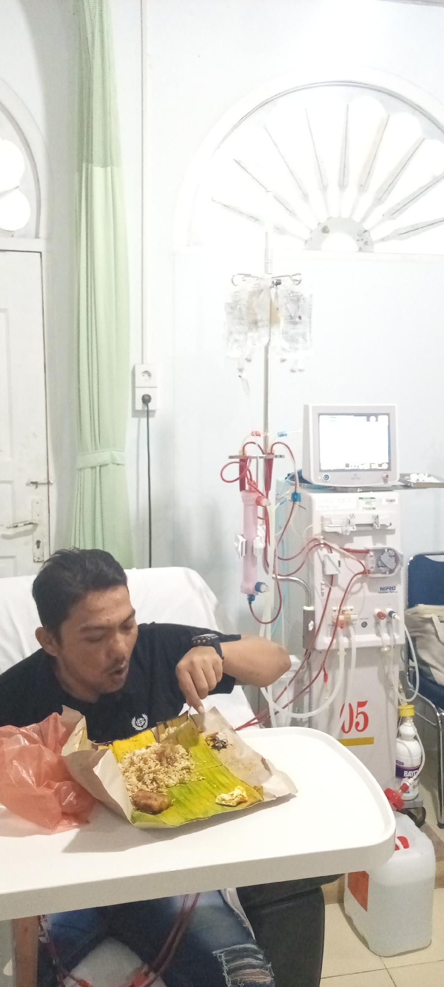 In a hospital room in Lhokseumawe, Indonesia, a man is sitting up in a hospital bed and eating from a plate covered with a banana leaf. He is wearing a black t-shirt and ripped blue jeans. To his right, a dialysis machine with multiple tubes and an IV stand with bags of fluid are visible. The room has a pale green curtain and a white electrical outlet.