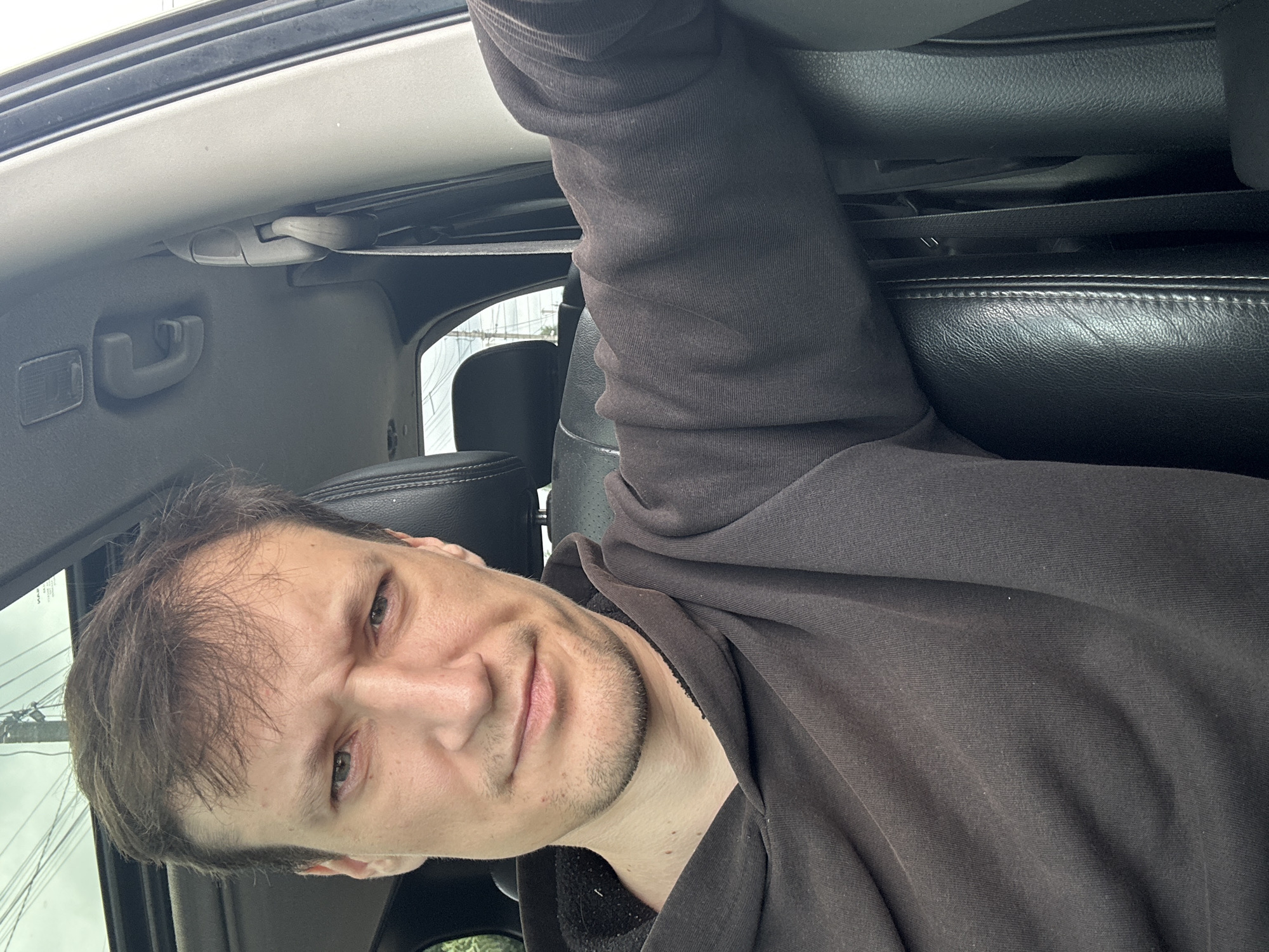 A man is sitting in the driver's seat of a car, looking at the camera. The man has light brown hair and a beard, and is wearing a dark brown hooded sweatshirt. The interior of the car is visible, including a black leather seat and a portion of the dashboard. The car appears to be parked, possibly in a residential area, as evidenced by the power lines and trees visible through the windshield. The image was taken in Makiv, Ukraine.