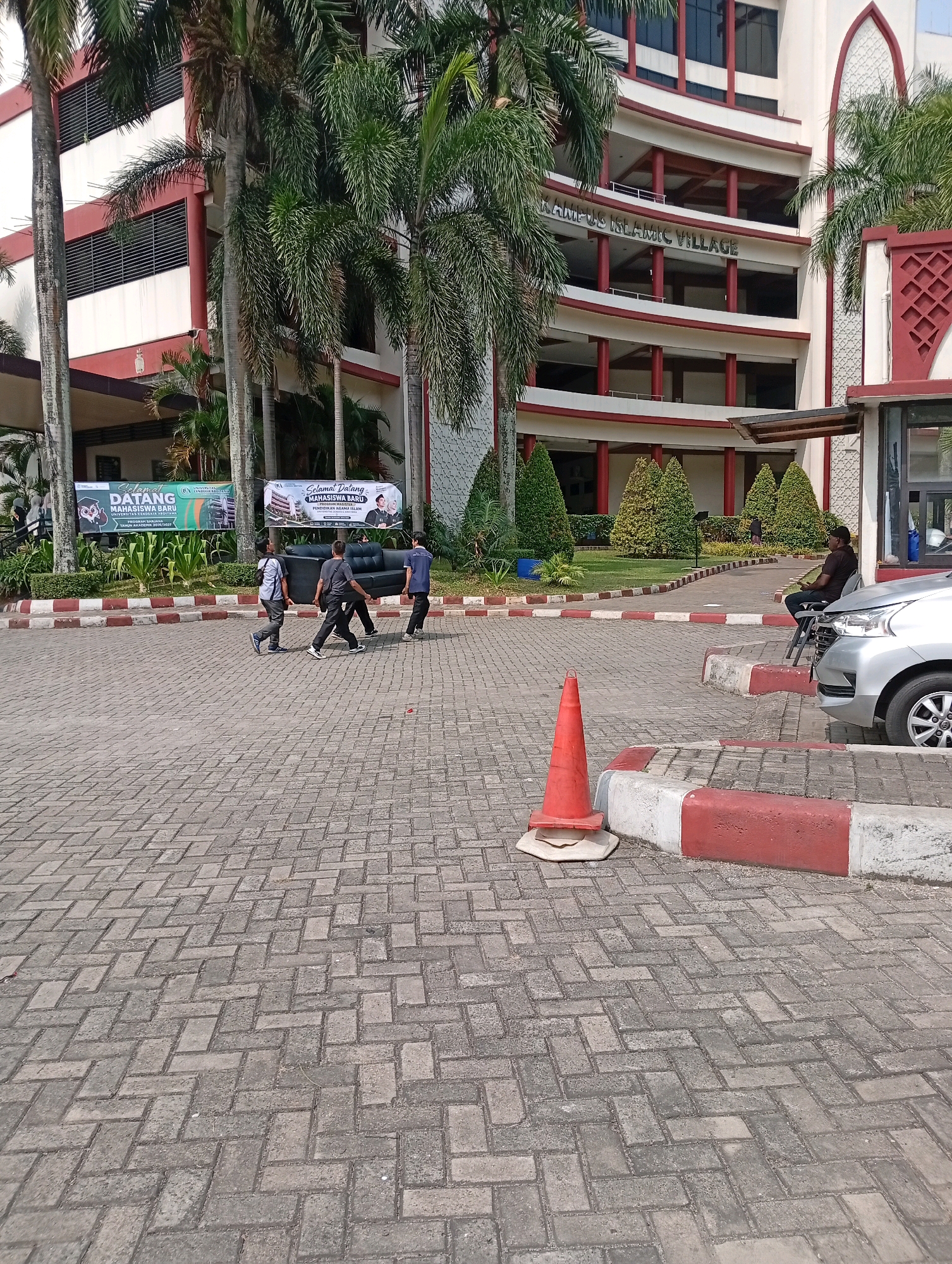 In the city of Kelapa Dua, Indonesia, at a campus identified by signage as "KAMPUS ISLAMIC VILLAGE", three men are observed carrying a dark gray couch across a paved area. The men are dressed casually: one in a light blue shirt and dark pants, another in a gray t-shirt and dark pants, and the third in a dark blue t-shirt and dark pants. They are walking from left to right in the frame. To the right of the men, a person is seated on a dark chair next to a silver car, holding a dark object, possibly a phone. In the foreground, an orange traffic cone is placed on a white and red striped curb. Behind the men and the traffic cone is a building with multiple stories, featuring red and white exterior elements and rows of windows. Several palm trees are visible in front of the building, along with manicured bushes and trees. Banners are also displayed on the building, one with the text "SELAMAT DATANG MAHASISWA BARU". The ground is paved with interlocking bricks. The lighting suggests it is daytime.