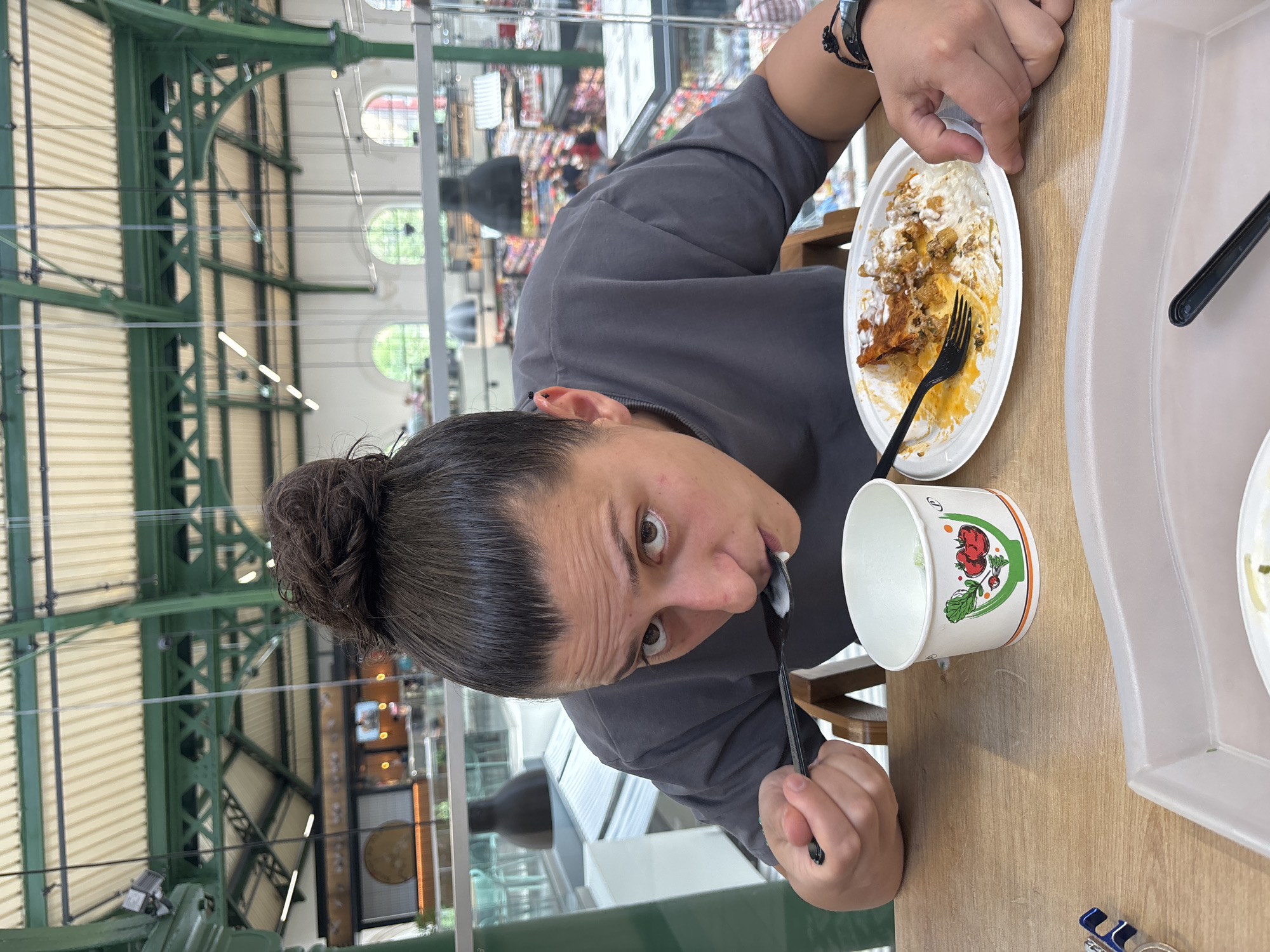 The image captures a person eating at a food stall in the Central Market Hall (Halite) in Sofia, Bulgaria. The person, with their hair pulled back in a bun, is leaning forward, holding a spoon and bringing it towards their mouth, actively eating. A partially eaten meal is on a white plate in front of them, with a fork beside it. To their left is a small paper cup with a drawing of tomatoes and lettuce on it. The background reveals the interior of the market hall, characterized by its distinctive green metal beams supporting a ribbed ceiling, with shoppers and market stalls visible in the distance. The lighting suggests it is daytime.