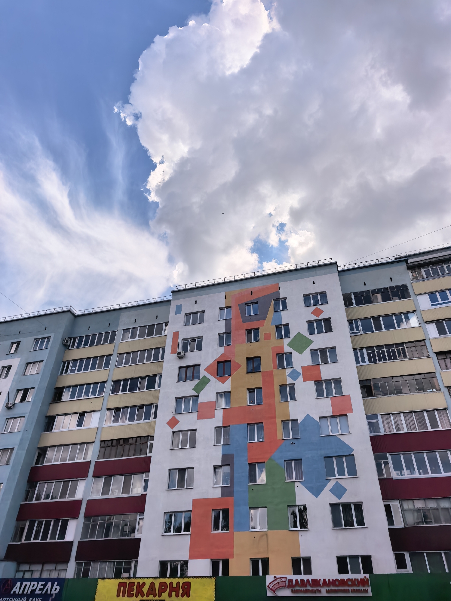 This is a low-angle, full-shot photograph of a brightly painted apartment building in Sterlitamak, Russia. The building is tall and appears to be made of concrete with multiple stories. The facade of the building is divided into sections of various colors, with a large, abstract geometric mural dominating the central portion. The mural features a pattern of squares and diamonds in vibrant hues of orange, red, yellow, blue, and green, set against a white background. The rest of the building is painted in more muted tones of light blue, yellow, and dark red. Numerous windows with white frames are visible across the entire structure, reflecting the sky and surrounding environment.
The sky above is a mix of bright blue and dramatic white clouds, with some wispy cirrus clouds stretching across the upper left quadrant and larger, more textured cumulus clouds on the right. The sunlight suggests it is daytime.
At the base of the building, a row of shops is visible, with signs in Russian. One prominent sign in yellow reads "ПЕКАРНЯ" (Bakery). Another sign to the left appears to say "АПРЕЛЬ" (April). A third sign on the right displays a red and white logo and text.
There are no people clearly visible in the image. The scene depicts an urban environment with a focus on the architectural and artistic elements of the apartment building. The overall impression is one of a modern, perhaps redeveloped, residential area.