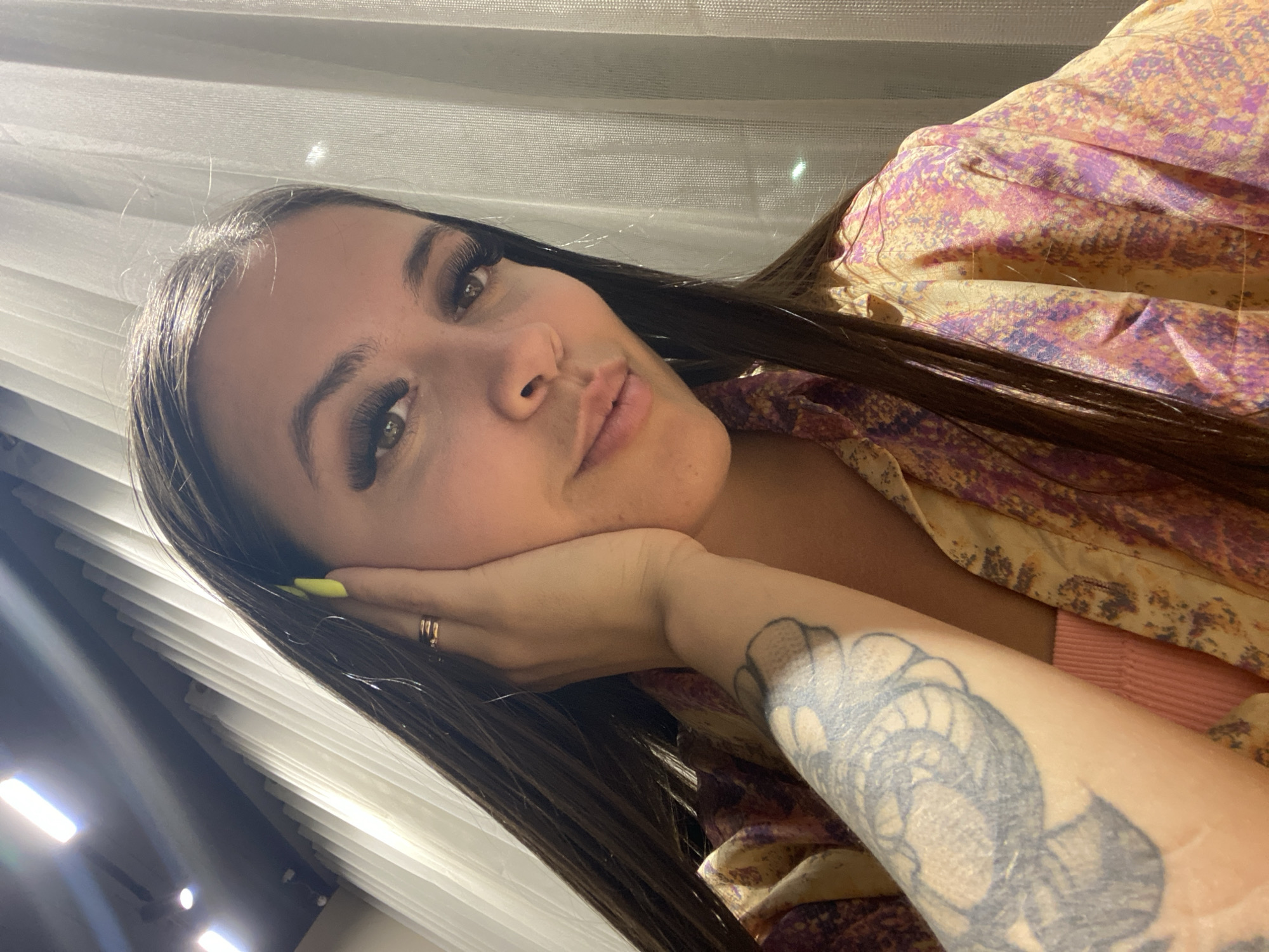 The media file shows a close-up, tilted selfie of a young woman resting her head on her hand. She has long, dark brown hair, prominent eyelashes, and is wearing a patterned top with shades of yellow, pink, and purple. A large tattoo is visible on her arm. She is wearing a ring on her left hand, and her fingernails are painted neon yellow. The background appears to be an indoor setting with white blinds and some lighting elements visible on the left side, suggesting it could be daytime. The woman's expression appears relaxed and perhaps pensive. There is no visible text in the image.