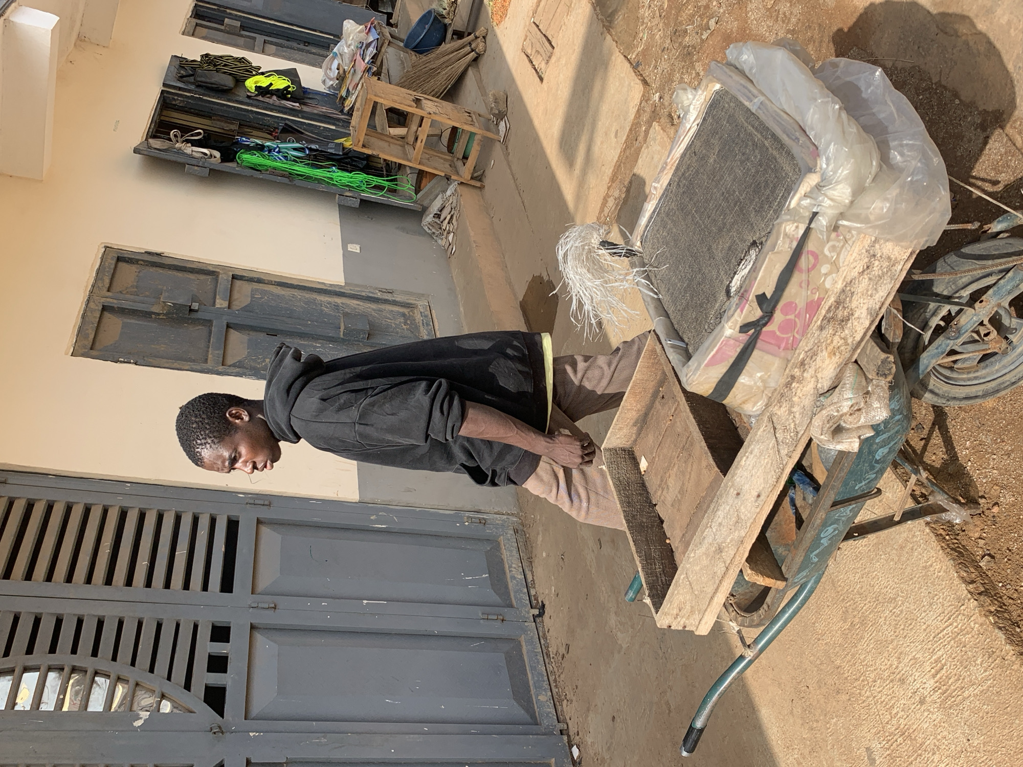 In Jos, Nigeria, a young man wearing a black hoodie and tan pants walks alongside a wheelbarrow loaded with goods. The wheelbarrow contains a wooden crate filled with smaller wooden objects, and on top, there's a larger, flat item wrapped in plastic sheeting, secured with black straps. The man is positioned on the left side of the frame, facing right in profile. Behind him, a building with light-colored walls features two large, weathered grey doors with horizontal slats. To the right, another doorway is visible, cluttered with various items including green ropes, a wooden ladder, brooms, and what appears to be a blue bucket. The ground is a mix of dirt and concrete. The scene suggests a market or a service area within a neighborhood.
