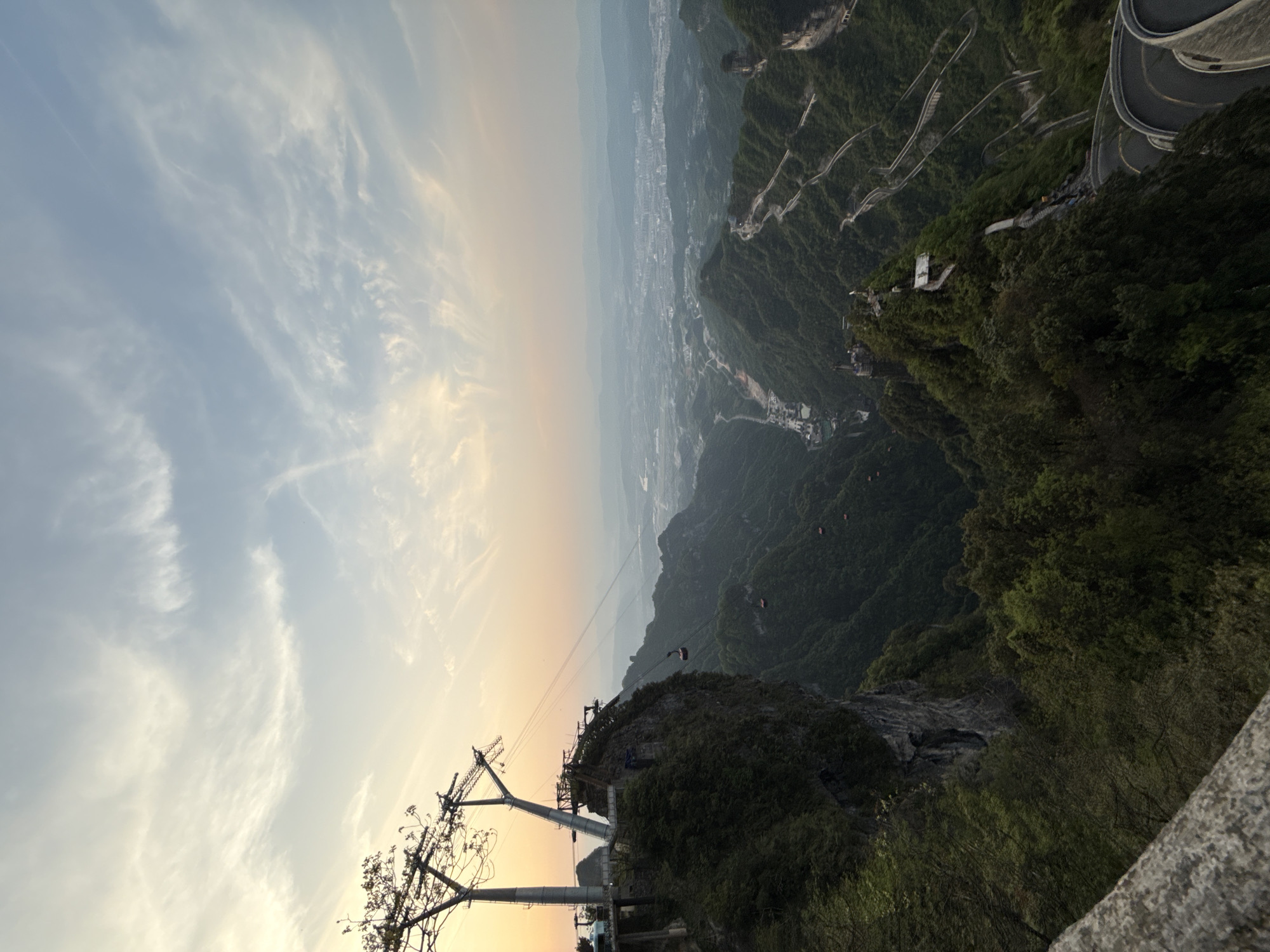 The image captures a landscape scene in Zhangjiajie, China, likely taken from a high vantage point, possibly a mountain or peak. The image is rotated.
Sky: The sky is partially visible, featuring a mix of light blue and white clouds, with a hint of yellow or orange near the horizon, suggesting sunrise or sunset.
Landscape: The landscape is dominated by mountains covered in lush greenery, and a distant city visible in the background. There are winding roads and paths carved into the mountain slopes.
Infrastructure: In the foreground, there is a cable car system visible, featuring support structures and a cable. A cable car is visible on the wire above a cliff.
Details: The image appears to have been taken during a clear day, and the lighting is soft.
Overall, the image portrays a scenic view of the mountainous terrain and infrastructure in Zhangjiajie, China, likely taken from a high-altitude perspective.