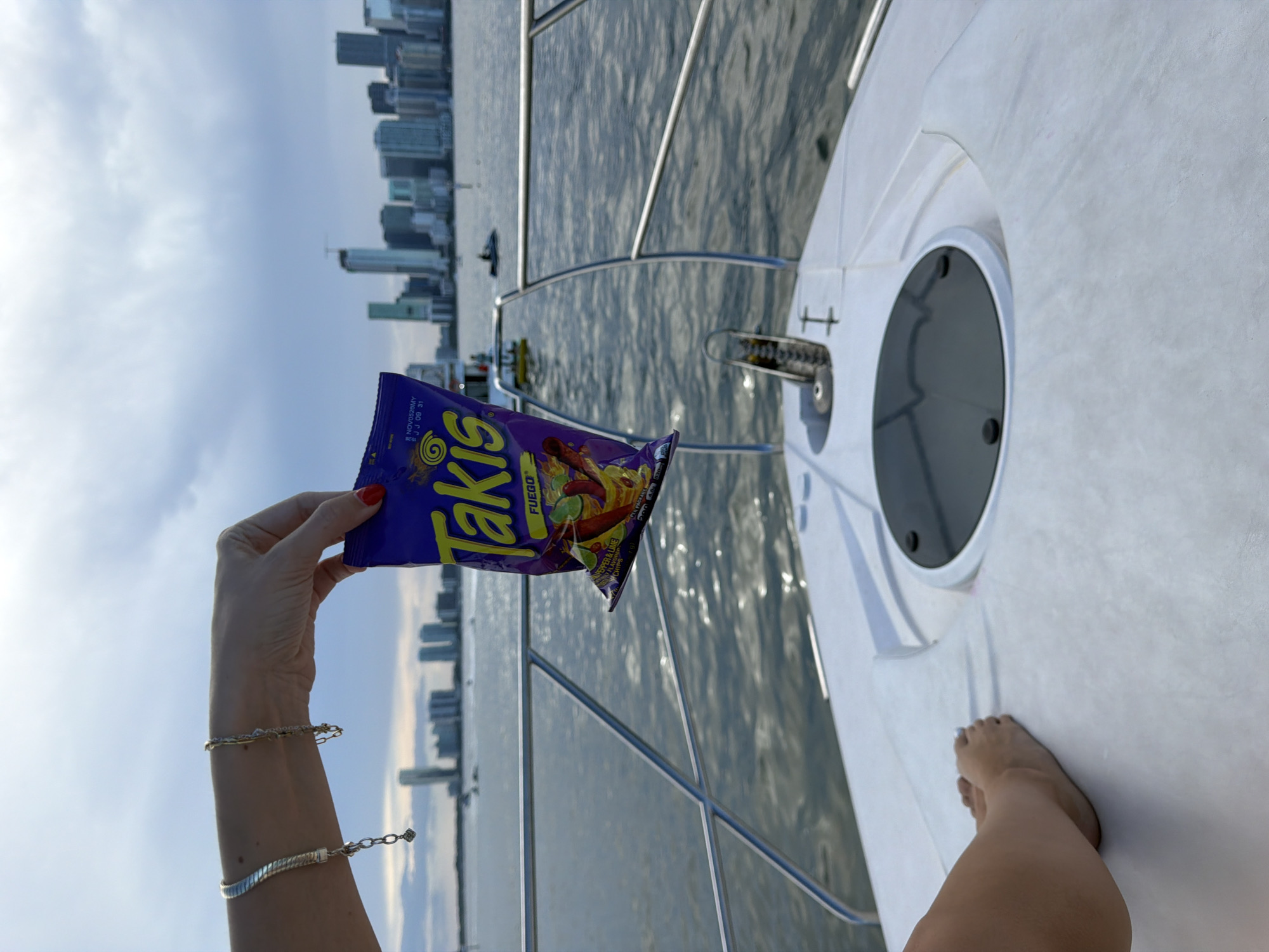 A woman's left arm, adorned with two bracelets, holds a purple bag of Takis Fuego chips. The woman's feet are visible on the deck of a white boat, which is on the water. In the background, the Miami skyline with its characteristic skyscrapers is visible under a cloudy sky. The water is choppy, reflecting the sky. The scene appears to be a recreational outing on the water with a view of the city.
