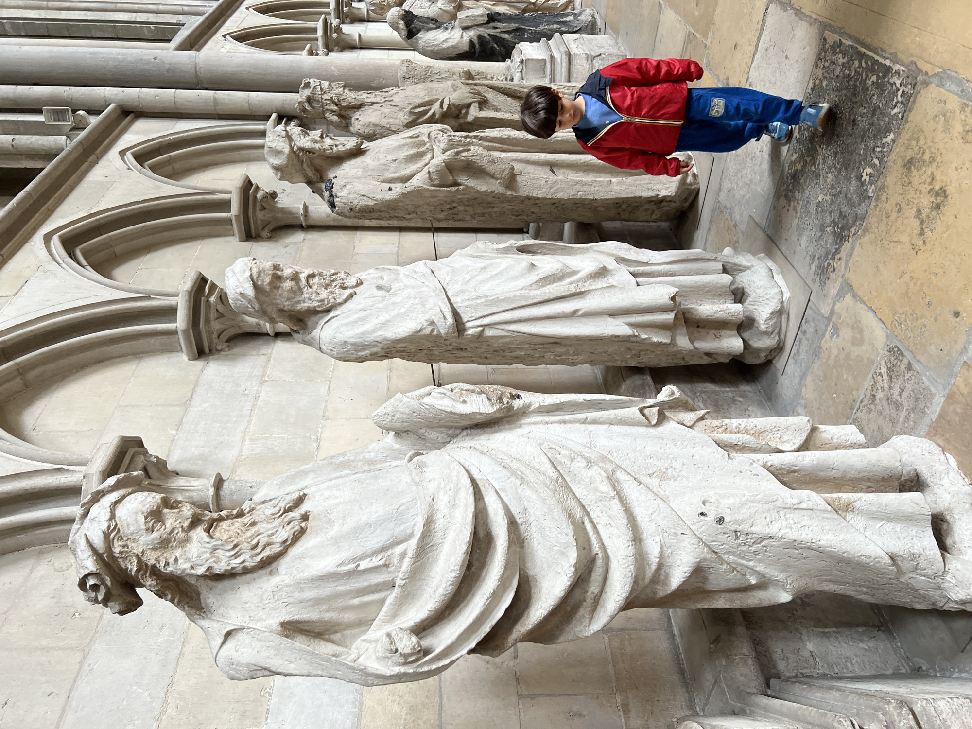 The image, if rotated 90 degrees clockwise, presents an indoor scene within a large, historic stone building, likely a cathedral or abbey. A young boy, wearing a red and blue jacket and blue trousers, stands on the right side of the frame, looking down at a row of ancient stone effigies or statues.
Several recumbent stone figures are arranged horizontally along a intricately carved stone wall. These figures are depicted in draped robes, with visible facial features and beards, suggesting they represent historical or religious individuals. Their surfaces appear aged and weathered, with intricate details in the carving of their garments and hair, as highlighted in the close-ups. The statues are made of a light-colored stone, possibly limestone or marble, and vary slightly in their state of preservation.
The architectural setting features prominent gothic arches, decorative pilasters, and finely worked stonework that forms the background. The floor is paved with large, irregular stone tiles, some lighter and some darker, contributing to the ancient atmosphere of the location.
The boy, with short dark hair, appears to be observing the stone figures with a sense of quiet curiosity. The lighting suggests it is daytime, illuminating the indoor space evenly. No discernible text is visible within the image. The scene conveys a sense of historical exploration and appreciation of ancient artistry and architecture.