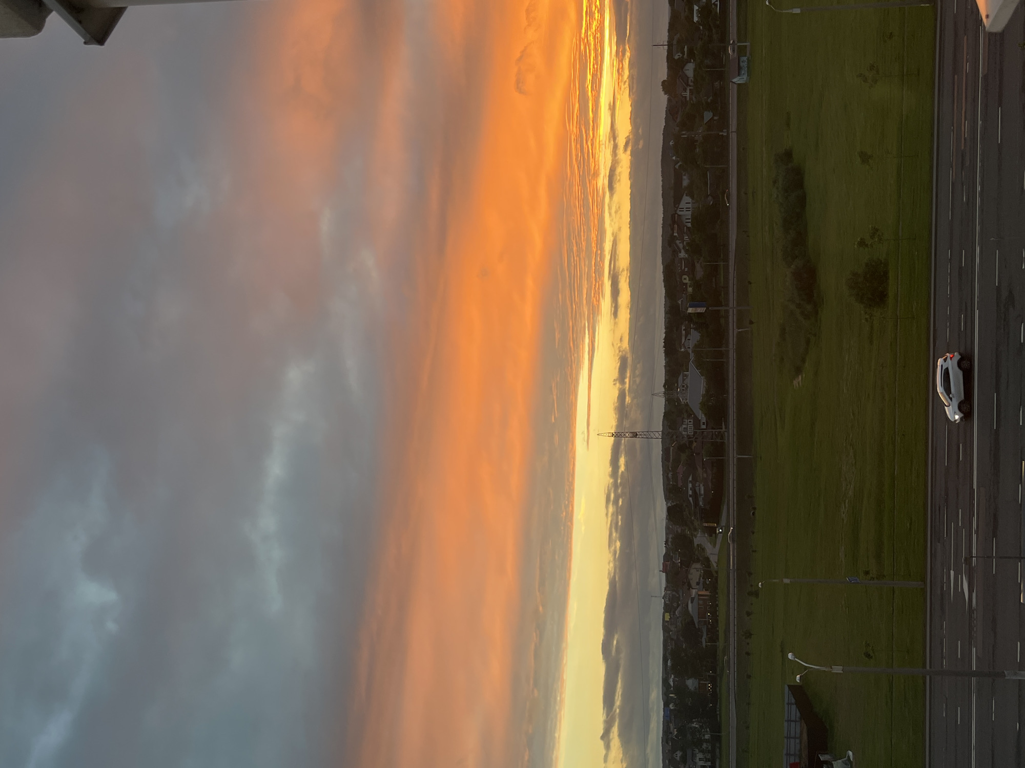 This image captures a sunset over a suburban area of Minsk, Belarus. The sky is filled with dramatic clouds, painted in hues of orange, yellow, and gray, with a touch of blue in the upper left quadrant. Below the vibrant sky, a landscape unfolds, featuring a mix of residential buildings, trees, and a grassy field. A road runs through the foreground, with a white car driving on it. In the midground, there appears to be a highway or a major road with some traffic, and further back, a collection of houses and structures indicative of a town or village. A tall transmission tower is visible in the distance, contributing to the urban or semi-urban skyline. The overall scene suggests a peaceful evening in a developing area on the outskirts of the city.