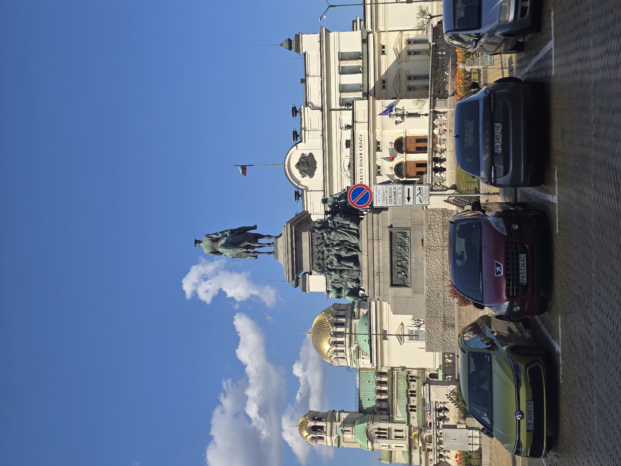 A bright, sunny street scene in Sofia, Bulgaria, captured during daylight hours, features a prominent equestrian statue and several notable architectural landmarks. The image is composed of a clear blue sky with scattered white clouds at the top, historical buildings in the midground, and parked cars along a cobblestone street in the foreground.
At the center-left, a grand equestrian statue, likely the Monument to the Tsar Liberator (Alexander II), stands on a tall, ornate pedestal adorned with sculptural reliefs. To the right of the statue, a large, light-colored Neoclassical building, identified as the National Assembly (Parliament) of Bulgaria, dominates the midground. It features several windows, a prominent facade with the Bulgarian national motto "СЪЕДИНЕНИЕТО ПРАВИ СИЛАТА" (Unity Makes Strength) inscribed above its main entrance, and is flanked by Bulgarian and European Union flags.
Further in the background and to the left, the distinctive golden domes and green roofs of the Alexander Nevsky Cathedral and possibly other churches are visible, adding to the city's iconic skyline.
Along the right side of the street, several cars are parked. A dark grey Skoda with license plate PB 3482 HE (Plovdiv region) is visible, along with a red Peugeot (PA 5136 KC, Pazardzhik region) and a lime green Volkswagen (CB 3600 CX, Sofia city). A red circular "no entry" sign and a white rectangular street sign are also present. The street sign indicates "СЛУЖЕБЕН АБОНАМЕНТ" (Official Subscription/Service Permit) parking with "2 бр." (2 pcs.) of spaces, along with arrows for direction and likely other parking information. The overall atmosphere is calm and clear, typical of a bright day in the city.