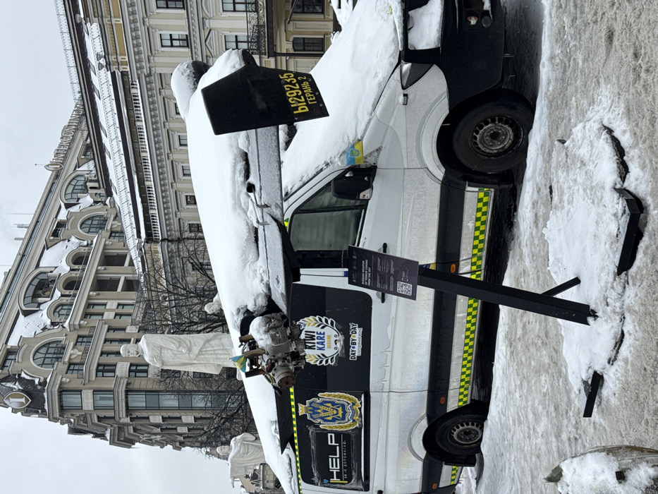 The image depicts a static outdoor display in Mykhailivska Square, Kyiv, Ukraine, amidst snowy conditions. A white Mercedes-Benz Sprinter van, partially covered in snow, serves as the primary display platform. Mounted atop the van's roof are significant remnants of what appears to be an uncrewed aerial vehicle (UAV) or missile. A large, dark-colored wing section, also snow-covered, extends from the passenger side roof. A black panel attached to the rear of this wing bears the yellow inscription "Ы29235 ГЕРАНЬ 2". Near the front of the van's roof, on the driver's side, a small, exposed engine assembly with metallic components is visible, likewise covered in snow.
The van's side features several decals: a Ukrainian heraldic-style emblem, a shield-shaped logo, "HELP S.O.S ДОПОМАГАЮ", and "KIWI KARE DAY-BY-DAY". A small Ukrainian flag sticker is affixed to the front of the hood. The lower perimeter of the van is lined with yellow and green checkerboard reflective tape.
In the mid-ground, behind the van, several large, white marble statues, depicting figures such as apostles or saints, stand on snow-covered pedestals. Beyond these statues, a multi-story building with ornate architecture is prominent, featuring "INTERCONTINENTAL" signage and a distinct "1" logo at its corner, identifying it as the InterContinental Kyiv hotel. The ground is covered in a layer of fresh snow. In the foreground, a black metal information stand with a sign displaying text, partially visible as "RUSSIAN WEAPON OF TERROR...", is erected on the snowy ground.