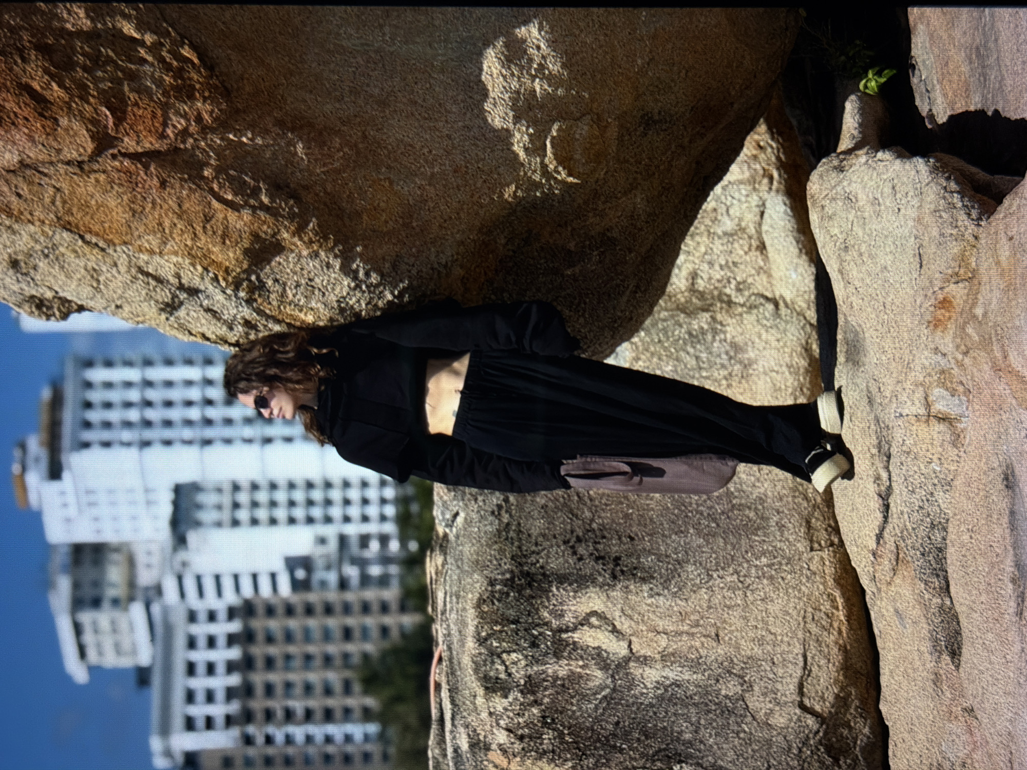 A full-body shot of a person, photographed outdoors, is presented against a backdrop showing a skyline of buildings. The subject is wearing black clothing, including a long-sleeved top and pants, complemented by white-soled shoes. The person appears to be positioned on rocky terrain. In the backdrop, a modern cityscape can be seen with the buildings stacked in the distant background. It is implied that this photo was taken in the city of Đà Nẵng, Vietnam.