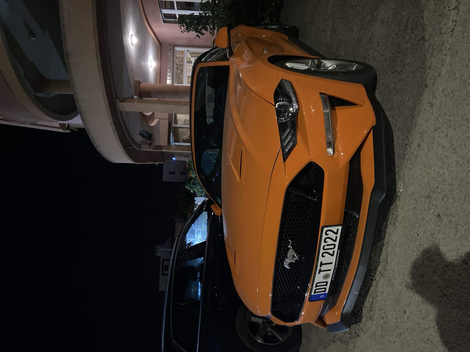 An eye-level, medium-shot image depicts an orange Ford Mustang parked outside a building in Bar, Montenegro. The car faces the right, taking up the right side of the frame. Its German license plate reads "DD TT 2022." The building has a curved architectural style, visible on the left side of the frame, along with a sign that says "RECEPTION." A dark SUV is parked behind the Mustang. The scene appears to be at night.