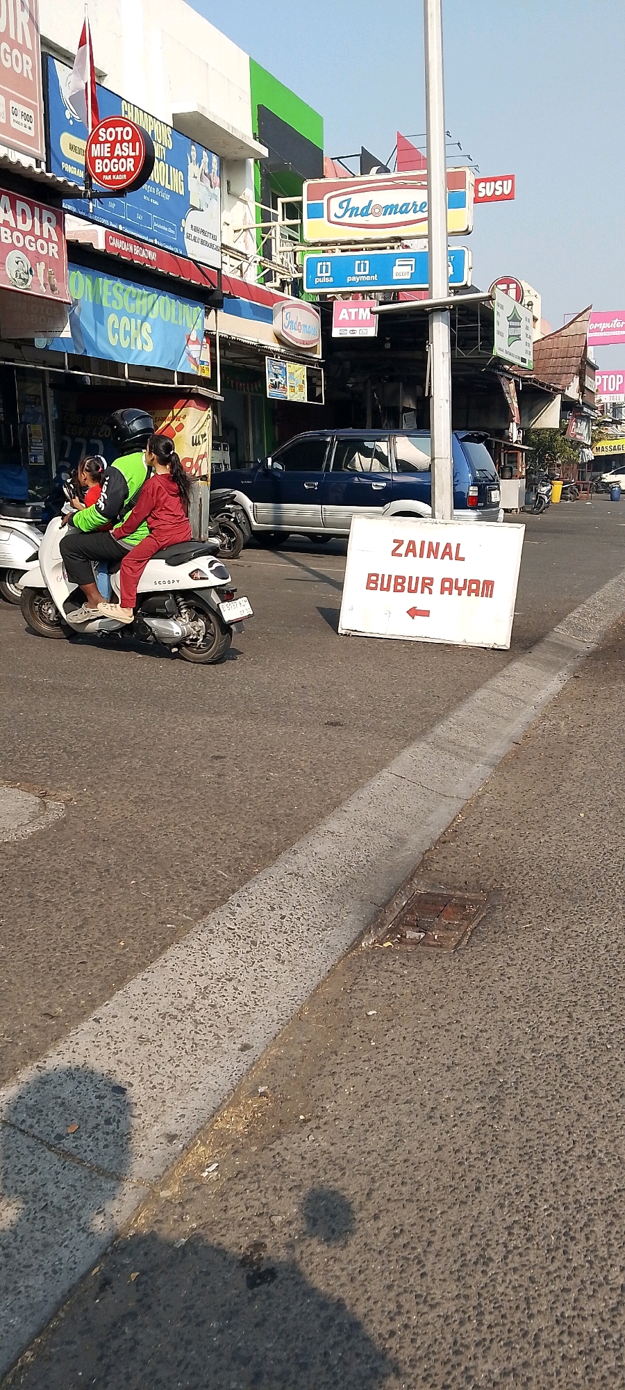 The image depicts a street scene in Cileungsi, Indonesia, under a clear sky. In the foreground, a white scooter is parked, and on the right side of the image, a white sign with red text reads "ZAINAL BUBUR AYAM" and features a red arrow pointing to the left. A white scooter carrying three people, a man and two young girls, is in motion, with the man operating the scooter and the girls seated behind him. All three individuals are wearing helmets. A dark blue SUV is visible in the background, parked or driving on the road. Numerous signs advertising various businesses, including "Indomaret," "SOTO MIE ASLI BOGOR," and "CHAMPIONS CITY SCHOOLING," are displayed on the buildings lining the street. The environment appears to be a commercial area with shops and a paved road.