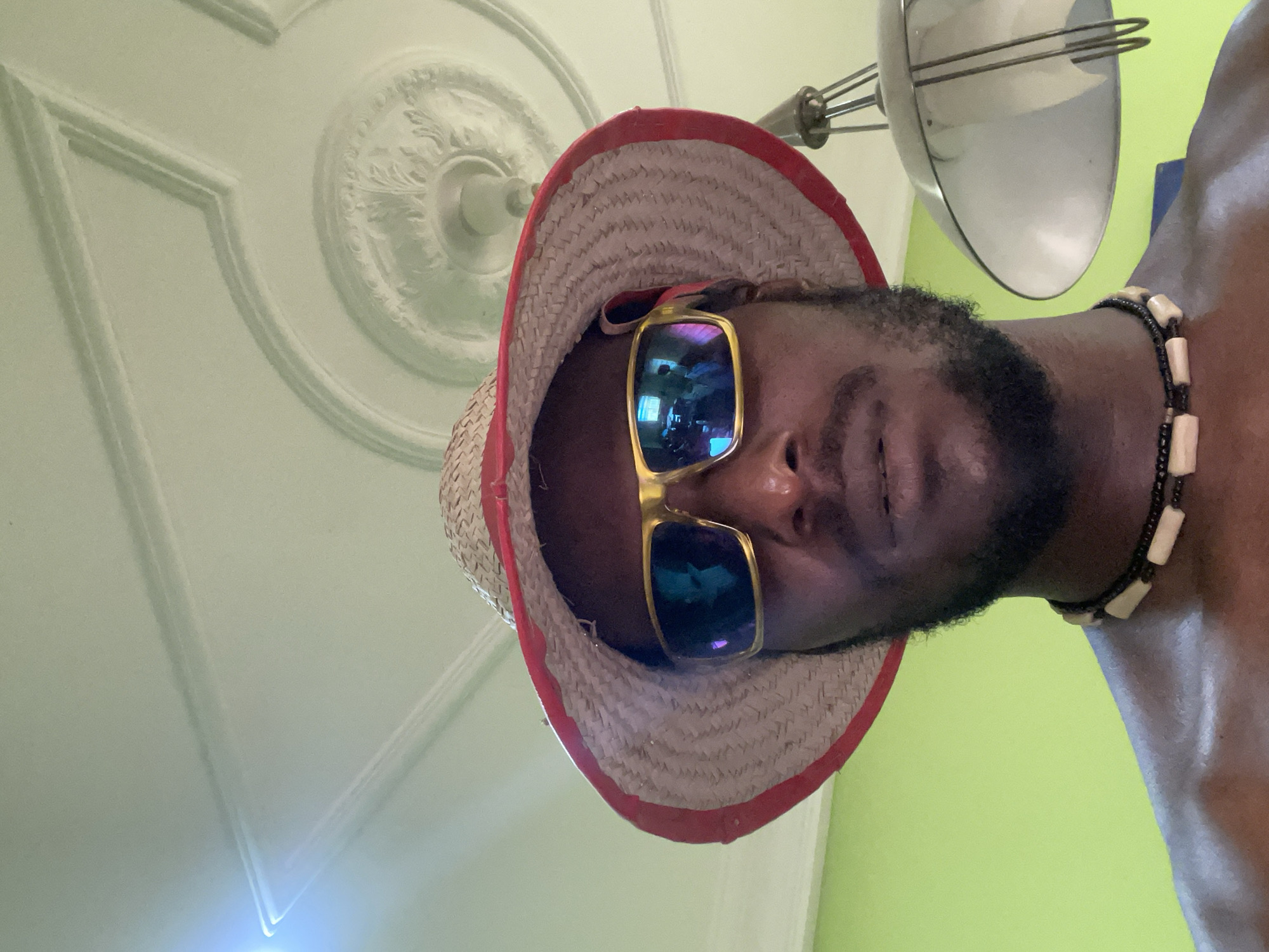 The image is a low-angle portrait of a person from the chest up, taken indoors. The individual, likely male, has dark skin, a neatly trimmed beard, and is looking slightly upwards towards the camera with a subtle smile. They are wearing a straw hat with a red brim and large, gold-framed sunglasses with highly reflective, iridescent lenses that obscure their eyes. A beaded necklace with black and off-white or bone-colored rectangular beads is visible around their neck.
The background features a light green ceiling with intricate white decorative molding, including a circular medallion and geometric panels. Part of a metallic light fixture or ceiling fan, resembling a whisk or a bowl-shaped shade with wire supports, hangs from the ceiling in the upper right. Below the ceiling, a vibrant lime green wall is visible. The perspective suggests the photo was taken from a low viewpoint, possibly a selfie. There is no visible text in the image. The setting is an interior space with distinct architectural details on the ceiling. While the location context is provided as Abuja, Nigeria, there are no specific visual cues within the image itself to confirm this geographical location. The time of day and weather are not discernible due to the indoor setting.