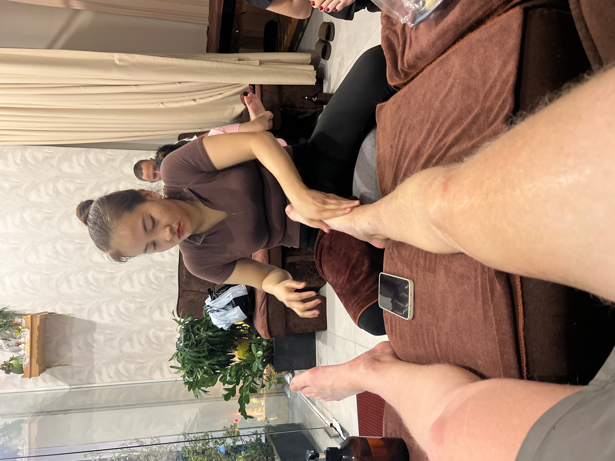 This image captures an indoor scene, likely within a massage parlor or spa in Bac Nha Trang Ward, Vietnam. The central activity depicted is a foot and leg massage.
In the foreground, a female massage therapist, wearing a brown short-sleeved top and dark pants, is intently massaging the lower leg and foot of a person whose fair-skinned leg and arm are prominently visible. Her hair is neatly tied back. The person receiving the massage is seated comfortably on a brown upholstered couch, with a white smartphone placed nearby on the upholstery. A brown pump bottle, likely containing massage oil, is visible in the bottom foreground.
In the background, other clients and staff are present. Another set of feet with red-painted toenails can be seen being massaged by someone out of frame, indicating multiple massage stations. A man's face is partially visible behind the main therapist, looking towards the viewer.
The setting is furnished with brown upholstered couches. The decor includes light-colored curtains, patterned white wallpaper, and green potted plants, contributing to a tranquil atmosphere. A large glass partition or window separates the interior from what appears to be an outdoor area with lush greenery. The lighting suggests it is either daytime or early evening. No visible text is discernible in the image.