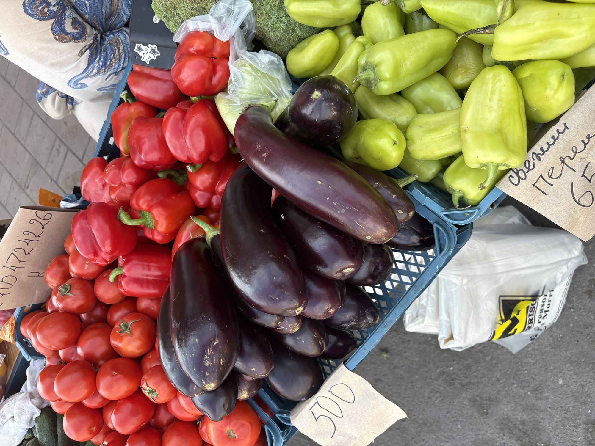 A close-up, overhead shot shows a vibrant outdoor market stall displaying an assortment of fresh produce. In the foreground, piles of dark purple eggplants are stacked high in a blue plastic crate. Next to them, a generous amount of bright green bell peppers are also arranged in a blue crate. Behind the eggplants, a colorful mix of ripe red tomatoes and red bell peppers are visible. A small sign with "500" written on it is tucked into the eggplants. Further back, a partially visible person is seated, their patterned clothing suggesting a traditional or decorative style. The ground appears to be paved, possibly asphalt, indicating an urban outdoor setting. Signs with handwritten text in Cyrillic are present, suggesting the location is likely in a Russian-speaking region or country, such as Astana, Kazakhstan, where such markets are common. The overall impression is of a bustling, fresh food market.