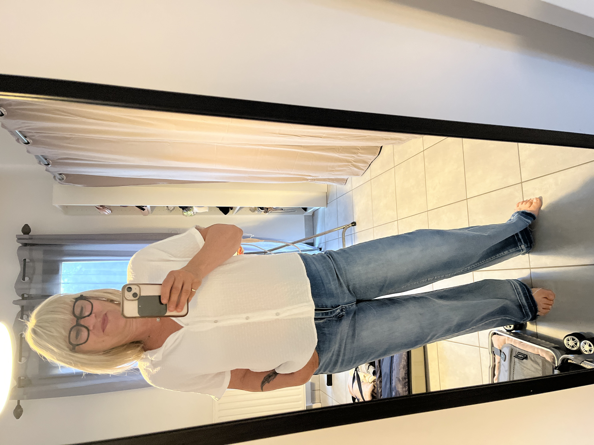 A middle-aged woman with blonde hair and glasses is taking a full-body mirror selfie in an indoor environment, likely a bedroom or dressing area. She is wearing a white button-down shirt and light blue jeans. The reflection in the mirror reveals a tiled floor, a suitcase with wheels, curtains, and a shelf with various items on it. The scene suggests a casual, personal setting. The location in Chambœuf, France is inferred.