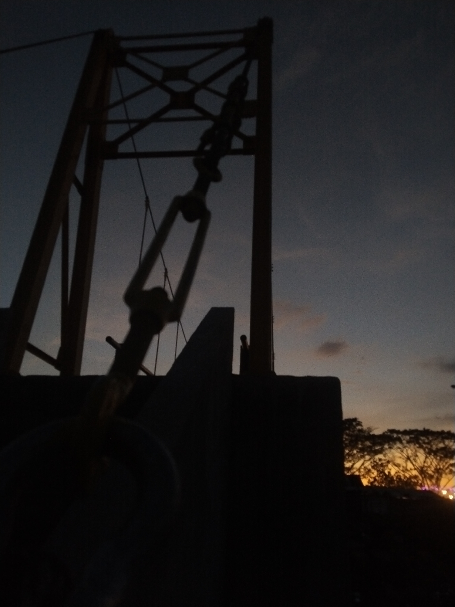 The image depicts a suspension bridge in Lhokseumawe, Indonesia, taken at dusk or dawn. The sky transitions from dark blue to orange near the horizon. The silhouette of a person standing on the bridge is visible. Trees and some scattered lights are visible in the background. The bridge structure and a tensioning device are prominently in the foreground, appearing as dark silhouettes against the sky.