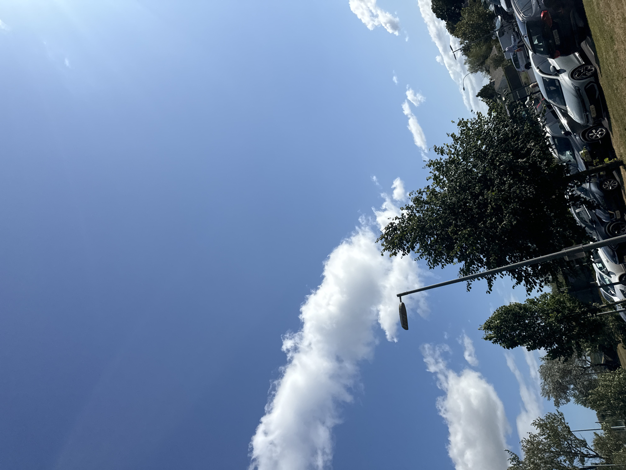 A bright, daytime, outdoor, medium-angle, eye-level shot shows a parking lot in Waterford, Ireland, on a clear day with a few scattered white clouds. The sky takes up most of the frame and is a brilliant blue. A long, thin white cloud stretches across the upper left quadrant of the image. In the lower right quadrant, parked cars are visible, including a dark-colored SUV and a white sedan with a license plate reading "222-WX-8906". A tall, grey lamp post with a rectangular light fixture stands in the middle of the frame. Several trees with dark green foliage are scattered throughout the scene. The ground appears to be grass and asphalt. The overall impression is of a sunny day in an urban or suburban environment.