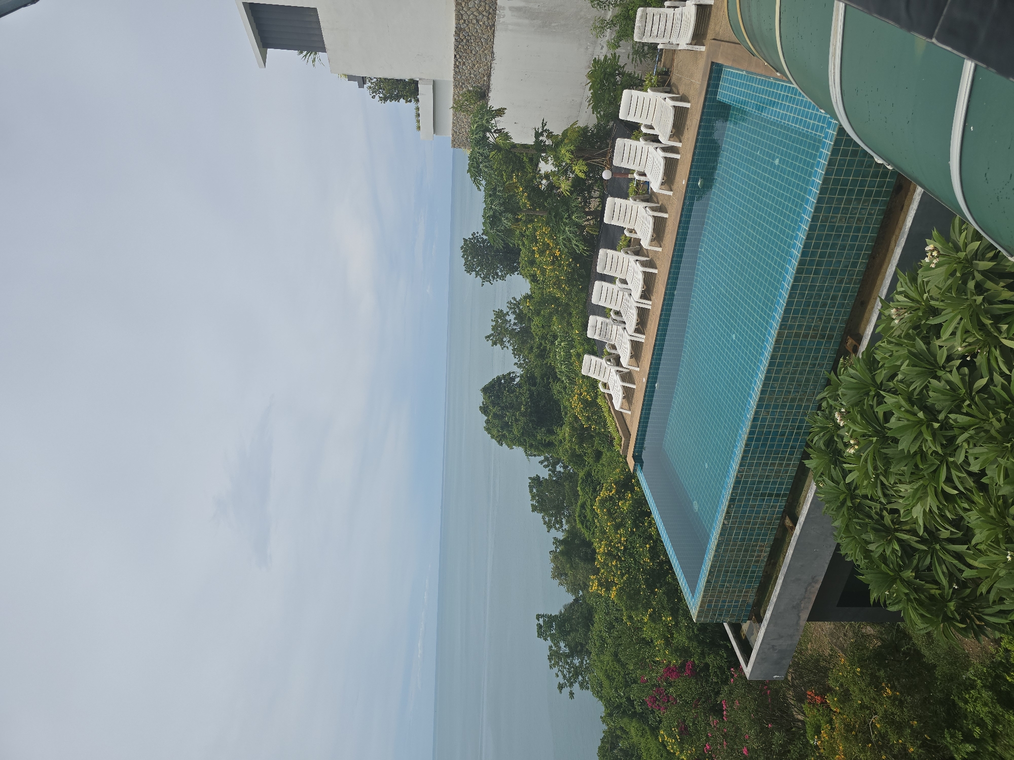 An aerial view captures a swimming pool overlooking a vast expanse of tranquil blue water and a lush green coastline. The infinity pool, lined with turquoise tiles, is empty, with several white lounge chairs neatly arranged along its edge. The surrounding landscape is a vibrant tapestry of dense, green foliage punctuated by splashes of pink and yellow flowers, hinting at a tropical setting. In the background, a modern, white building with stone accents rises above the greenery, its architecture suggesting a resort or hotel. The sky above is overcast, casting a soft, diffused light over the scene.