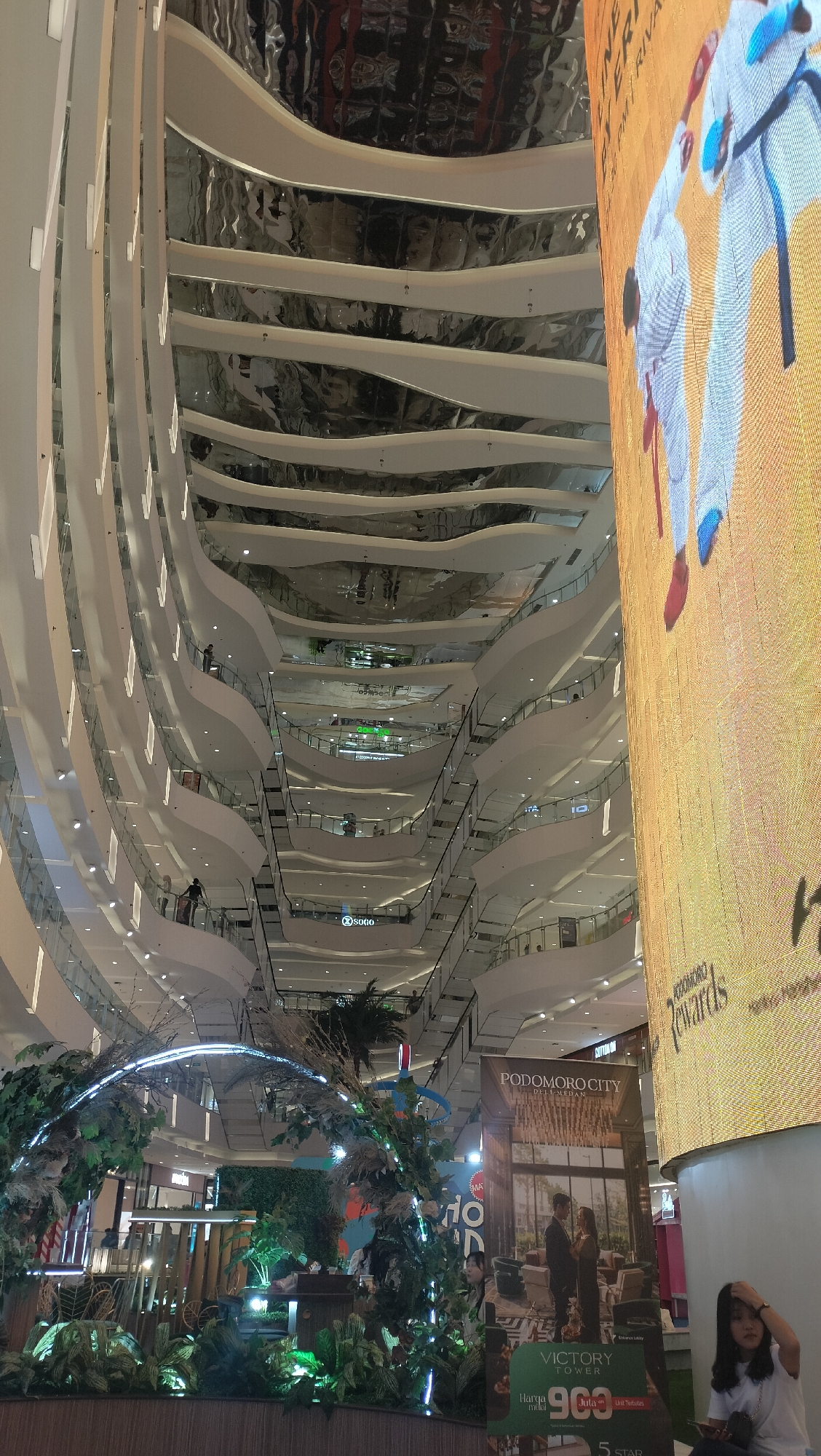 The image captures the interior of a multi-level shopping mall in the City of Medan, Indonesia. The architecture features a distinctive undulating design with multiple curved balconies and escalators visible. A large, illuminated digital screen dominates the right side of the frame, displaying an advertisement. On the left, a decorative archway adorned with greenery and lights is positioned in the foreground. Several individuals are dispersed throughout the mall, engaged in various activities such as walking and standing on the balconies. The overall environment is brightly lit, suggesting it's a public indoor space.