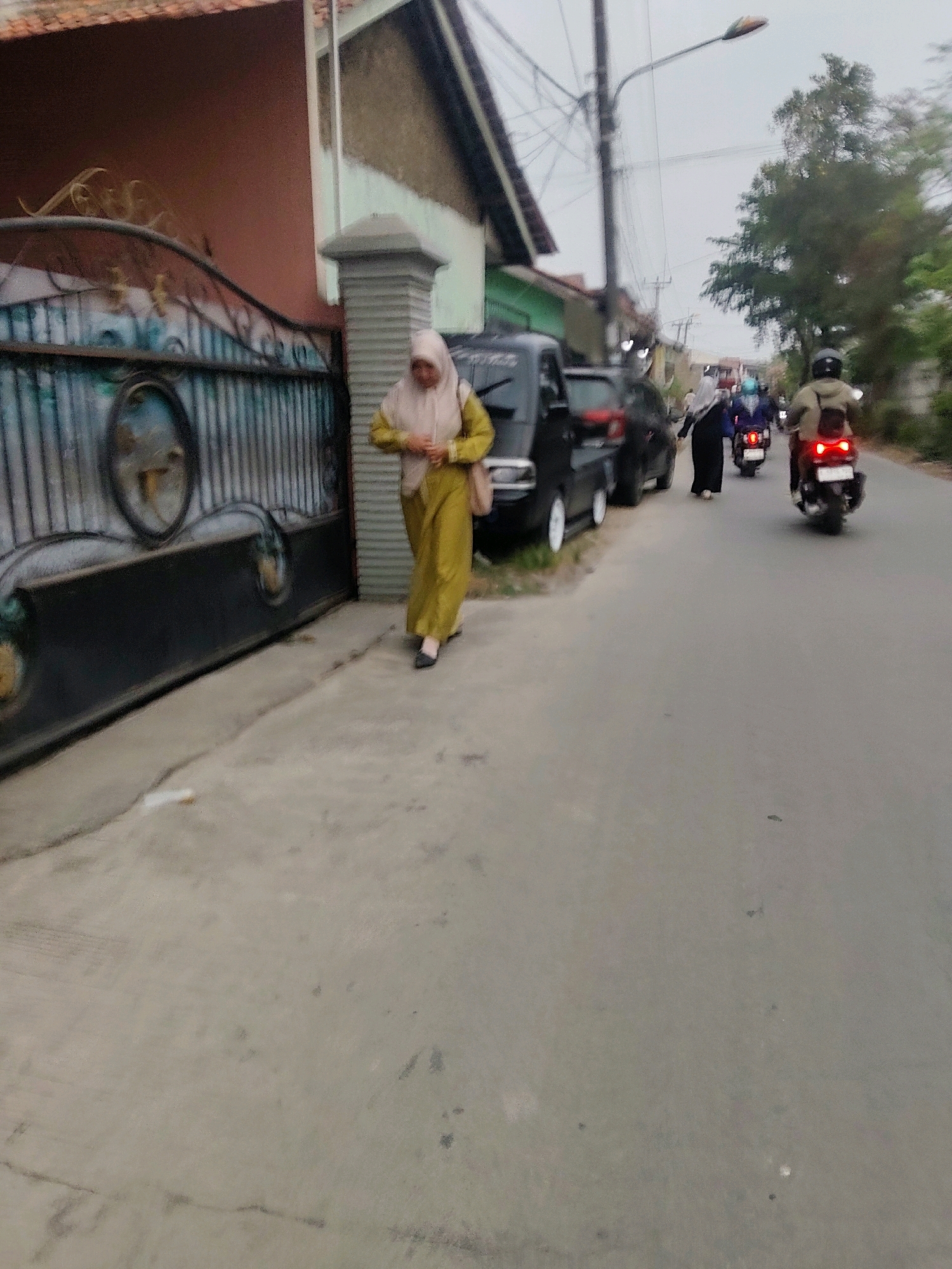 A woman in a yellow headscarf and matching outfit walks on a concrete sidewalk beside a dark metal gate. She is carrying a tan purse over her right shoulder. Behind her, a black van is parked on the side of a paved road. Further down the road, several motorcycles are visible, some with riders wearing helmets. The environment appears to be a street scene in Mekarmukti, Indonesia, with buildings and trees lining the road under an overcast sky.