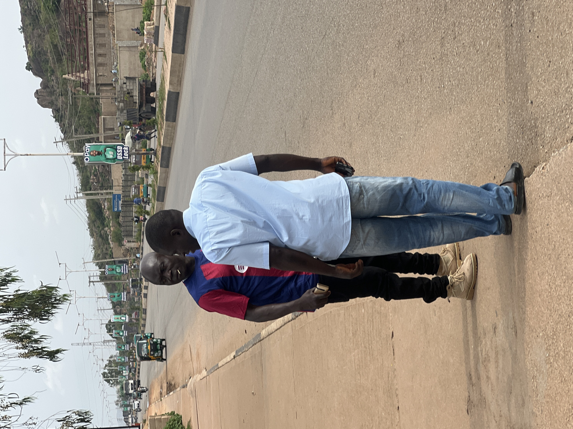Two men stand on the shoulder of a paved road in Gura Top, Nigeria. One man wears a light blue long-sleeved shirt, blue jeans, and black shoes. The other man wears a blue and red striped short-sleeved shirt, black pants, and tan boots. Both men have dark skin and short hair. They are holding phones. The road appears to be a busy street with vehicles and billboards in the background. The landscape includes a rocky hill or mountain and power lines.