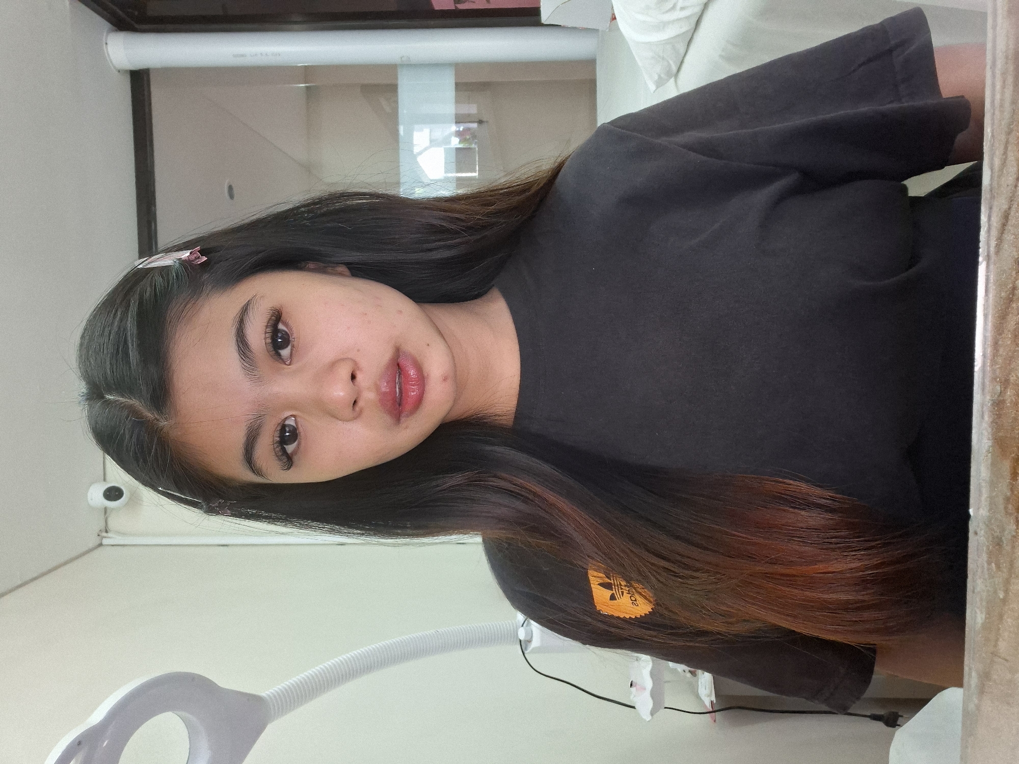A young woman with long, dark hair and subtle makeup is captured in a close-up selfie. She is wearing a dark t-shirt with an orange logo on the left chest. The background appears to be an indoor setting, possibly a room or a salon, with a white wall and a white lamp on the left. A security camera is visible on the wall behind her head. The overall lighting is soft, and the image is slightly grainy. This scene is not identifiable as being in Kuta, Indonesia.