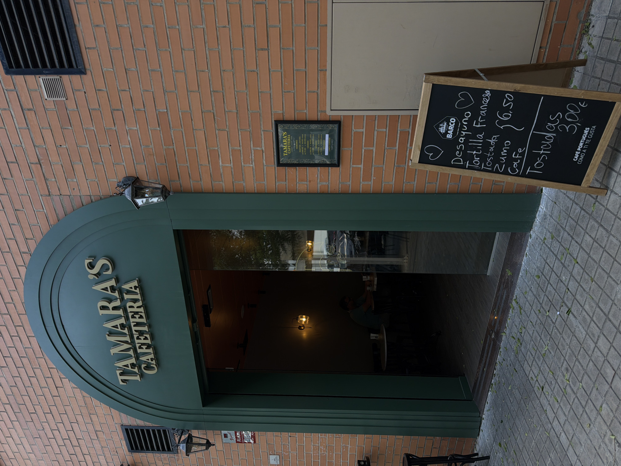 The image shows the entrance to "Tamara's Cafeteria" on a brick building. The cafeteria's name is prominently displayed in gold lettering above the arched doorway. To the left of the entrance, a vintage-style lantern hangs on the brick wall. Inside the cafeteria, visible through the open glass door, a person is seated at a table in a dimly lit interior with a hanging lamp. In front of the entrance, a wooden A-frame chalkboard displays a menu. The menu lists items like "Desayuno," "Tortilla Francesa," "Tostada," "Zumo," and "Cafe," with prices for some items. The exterior is paved with square tiles, and scattered leaves are visible on the ground. A framed schedule of the cafeteria's hours of operation is also affixed to the brick wall to the right of the entrance.