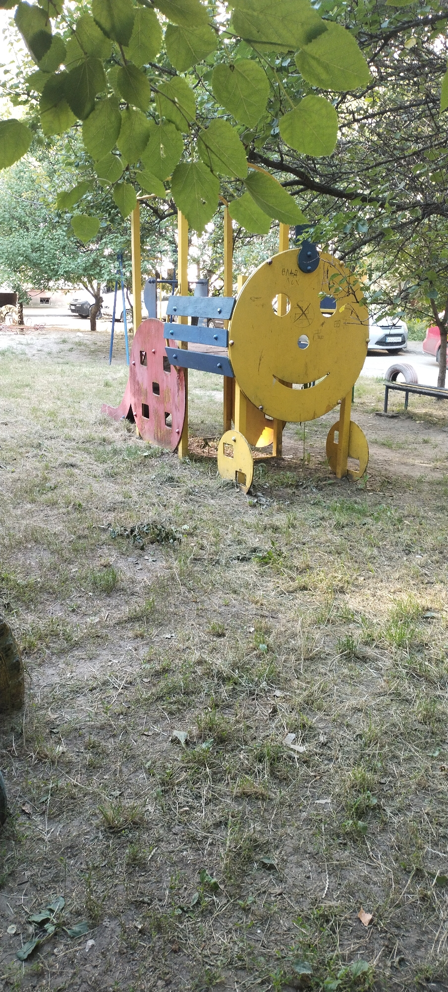 A yellow, circular playground structure with a smiling face, resembling a train car, is situated on a grassy area in Pisochyn, Ukraine. The structure features blue and gray slats forming a bench. A red, square-shaped component with openings is attached to the left side of the yellow structure. The ground is covered with dry grass and dirt, with some scattered leaves. Trees with green foliage are visible in the background, with a portion of a building and a car in the distance. The lighting suggests it is daytime.