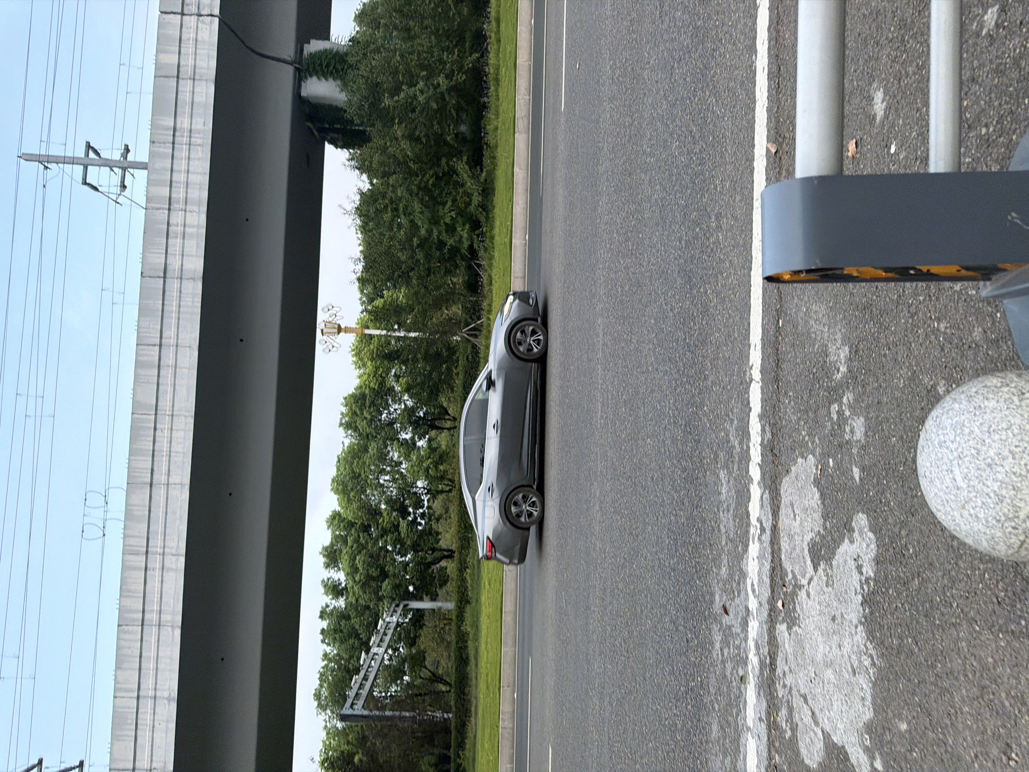 This is a side-view, medium-shot photograph of a grey sedan driving on a multi-lane asphalt road. The car is in motion, positioned in the middle lane, and appears to be traveling away from the viewer towards the right side of the frame. The road has white lane markings. In the background, there is a lush green area with trees and a significant elevated concrete structure, likely an overpass or bridge, with power lines running above it. The sky is overcast with some visible power poles and their associated infrastructure. The foreground shows the edge of the road with some white markings and a stone bollard. The scene is set in the city of Jiangjin, China, under a cloudy sky.