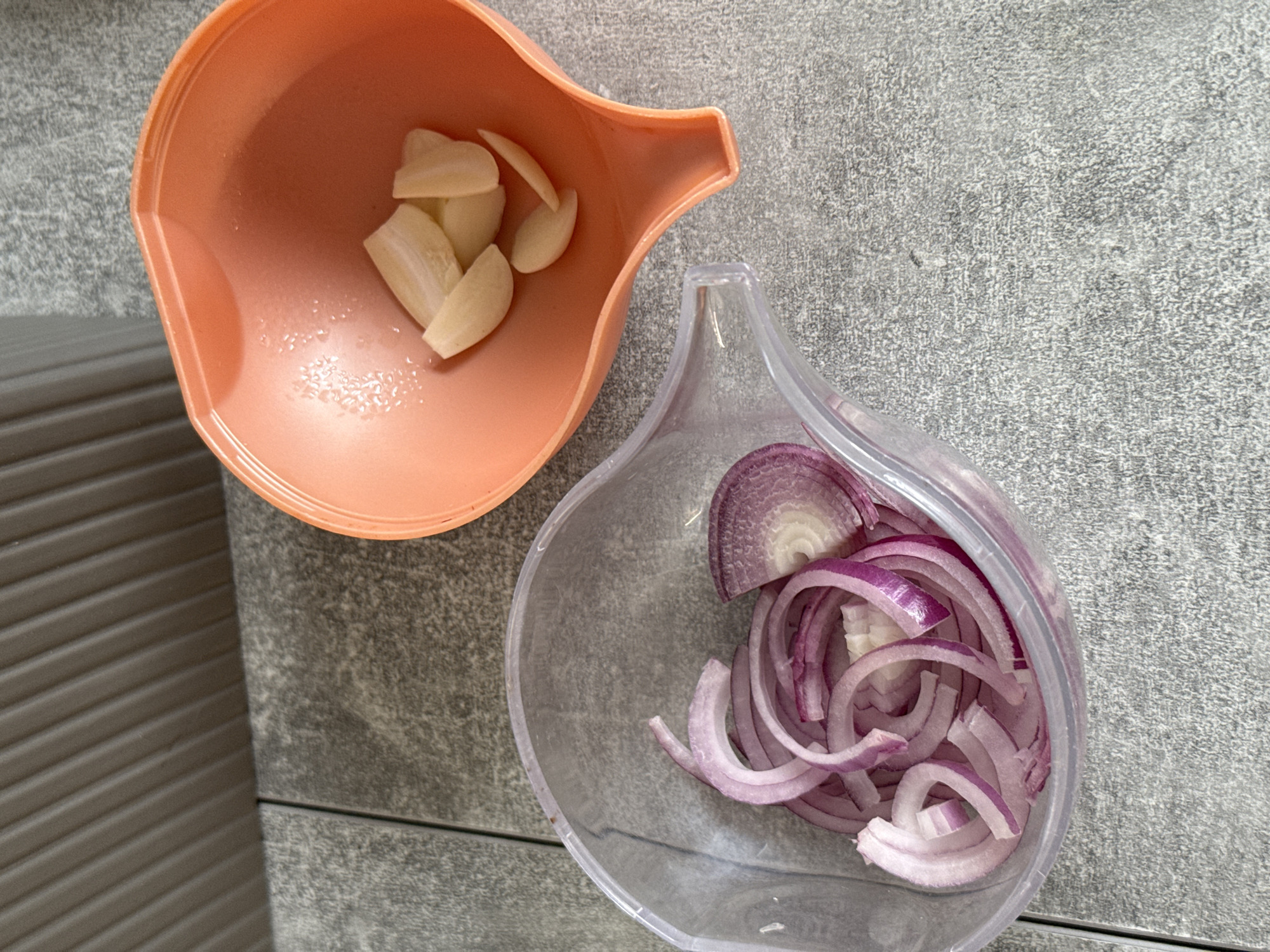 The image displays two plastic containers on a gray tiled surface. The container on the left is clear and filled with sliced red onions. The container on the right is orange and contains several cloves of peeled garlic. The background shows a wall with vertical gray ridges. There are no people or animals visible in the image. The scene appears to be in a kitchen or food preparation area. No specific location in Kraskovo, Russia can be determined from the image.