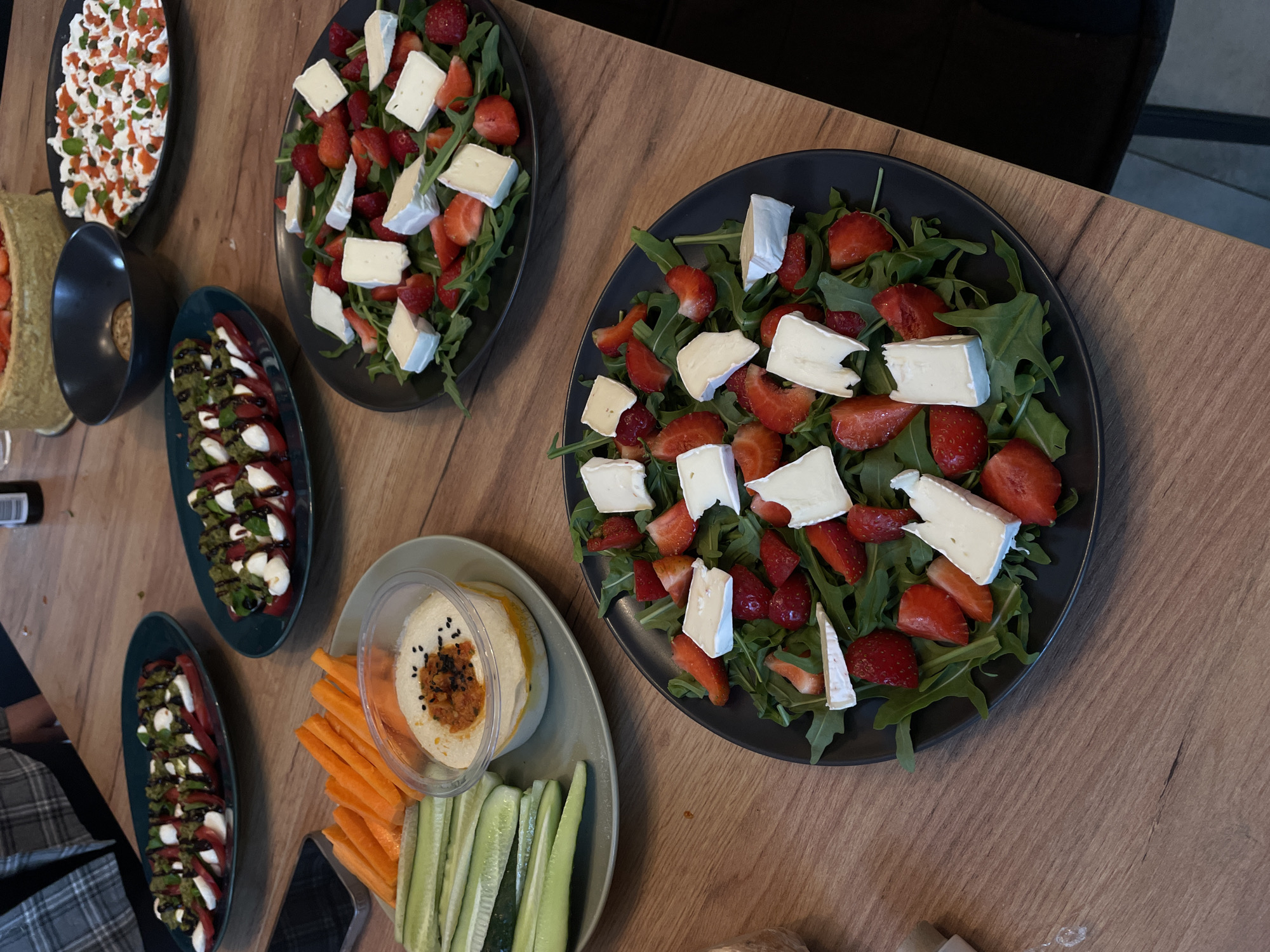 A close-up, eye-level shot captures a spread of appetizers on a wooden table. In the foreground, a dark plate holds a salad of arugula, sliced strawberries, and cubed cheese. To the left and slightly behind, another dark plate features sliced cucumbers and carrots served with a bowl of hummus topped with black seeds and a red garnish. Further back, two more dark plates display caprese salads with sliced tomatoes, mozzarella, and a green sauce. A dark bowl sits empty in the middle background, and a partially visible cake with strawberries is at the top. A person's arm and shoulder, wearing a plaid shirt, are visible on the left side of the frame, resting on the table. The setting appears to be indoors, likely a home or restaurant dining area, with no visible windows or distinct architectural features.