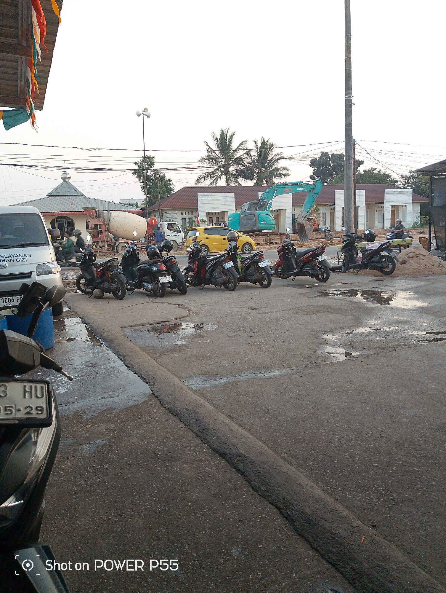The image shows a construction site in Batam, Indonesia, with a cement mixer truck, an excavator, and several motorcycles parked on a wet asphalt road. A building with a dome is visible in the background, possibly a mosque. There are also some residential buildings under construction. The scene suggests ongoing development or renovation work in the area. The lighting indicates it might be late afternoon or early morning. The overall impression is one of a busy urban environment undergoing construction.