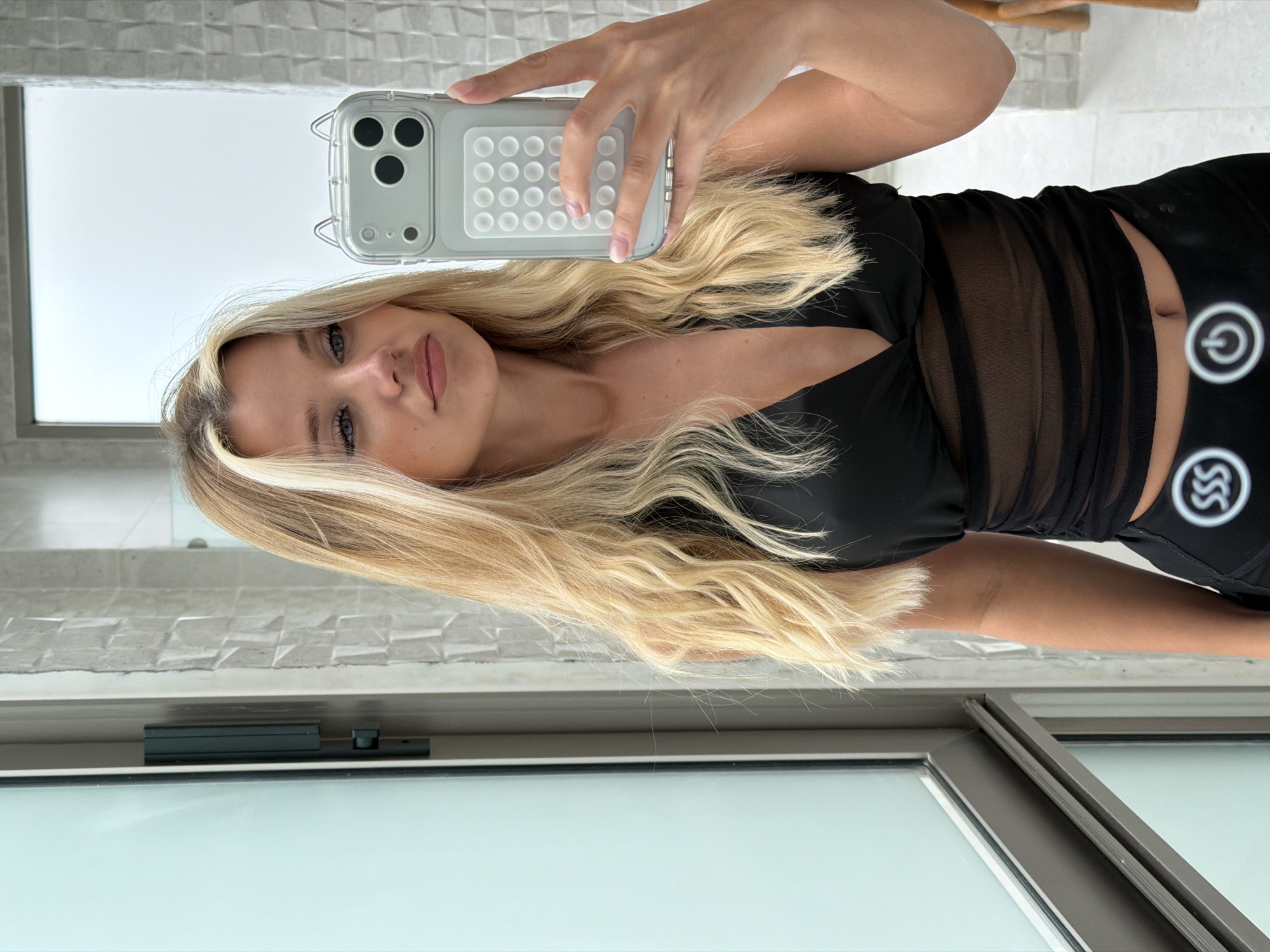A young woman with blonde hair is taking a mirror selfie in a bathroom. She is wearing a black, sheer top and black bottoms with a logo on the waistband. She is holding a phone in her right hand, which has a clear case with a grey grid pattern on the back. The bathroom has tiled walls and a large window. The image does not provide information about a specific location in Thep Krasatti, Thailand.
