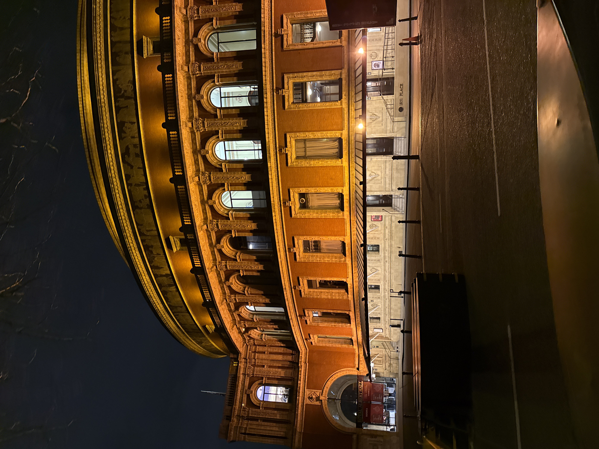 The image depicts the exterior of the Royal Albert Hall at night, located in Kensington Gore, London, United Kingdom. The large, circular, multi-level building, constructed of red brick, is brightly illuminated by warm, golden-yellow floodlights, highlighting its architectural details.
The structure features multiple tiers of arched and rectangular windows, some internally lit, with curtains visible in several lower-level windows. An ornate frieze depicting figures in bas-relief encircles the upper portion of the building. Decorative balustrades are visible on various levels. A prominent arched entrance on the left bears signage indicating "ROYAL ALBERT HALL" and "DOOR 6." Below a continuous, extended canopy, multiple ground-level entrances with steps are visible, and the text "1851 PLACE" is discernible on the facade near these entry points.
The immediate foreground contains a dark, indistinct rectangular object, potentially a barrier or bench. An asphalt road, appearing somewhat reflective, occupies the mid-ground, traversed by a white lane marking. Black bollards line the pavement edge, and a single orange traffic cone stands on the pavement to the right. The sky overhead is dark, with subtle patterns suggesting bare tree branches in the upper frame. No individuals or specific actions are present within the scene.