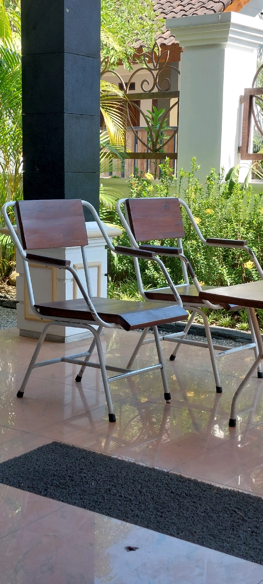 In a residential outdoor setting, possibly a porch or patio, two wooden-seated chairs with white metal frames are positioned on a tiled floor. The chairs are arranged side-by-side, facing slightly to the right. Behind them, lush green foliage, including palm fronds and smaller plants, suggests a tropical or subtropical environment. A decorative metal fence and a white pillar add architectural elements to the scene. The flooring is a light-colored tile with a glossy finish, reflecting the surrounding light and greenery. In the foreground, a dark grey mat is partially visible on the floor. The overall atmosphere is serene and suggests a place for relaxation.