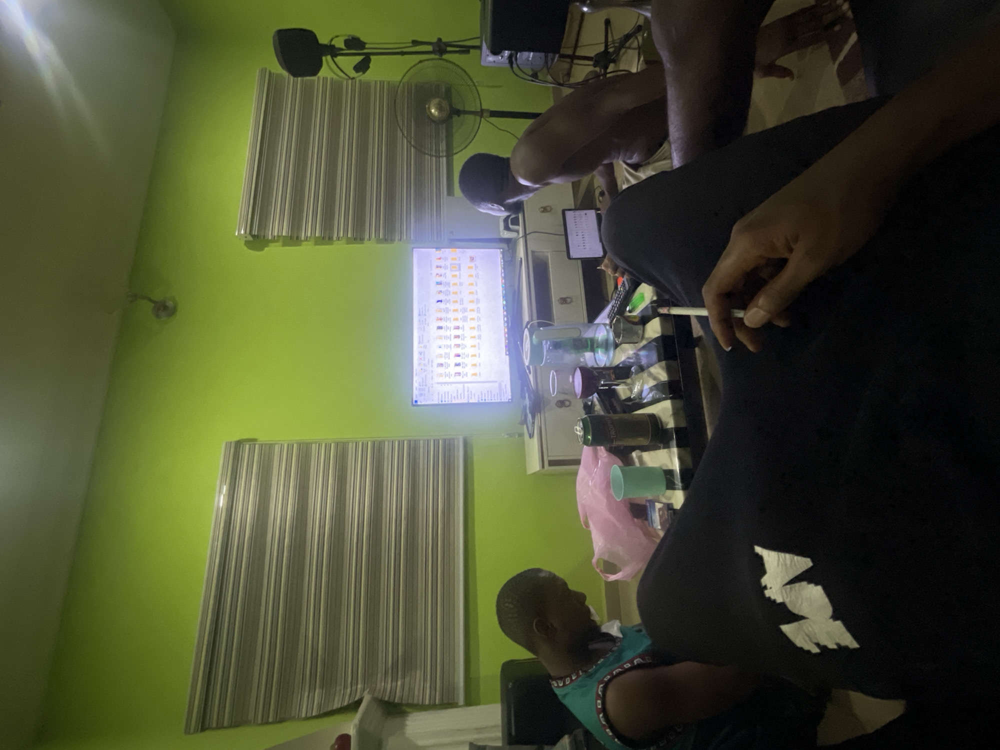 A brightly lit room with lime green walls. Two windows with closed Venetian blinds. Three men are present. One shirtless man is hunched over a table, looking at an open tablet. Another man is seated, partially obscured by the foreground figure, and is wearing a blue and green patterned tank top. The foreground man is wearing a dark colored shirt or jacket. This man is holding a white cigarette between his fingers. On the table are several drinks, including a can of beer and multiple clear plastic containers. A monitor displaying many small icons is also visible. There is a standing fan in the background. The location is most likely a private residence. The city and country are provided.