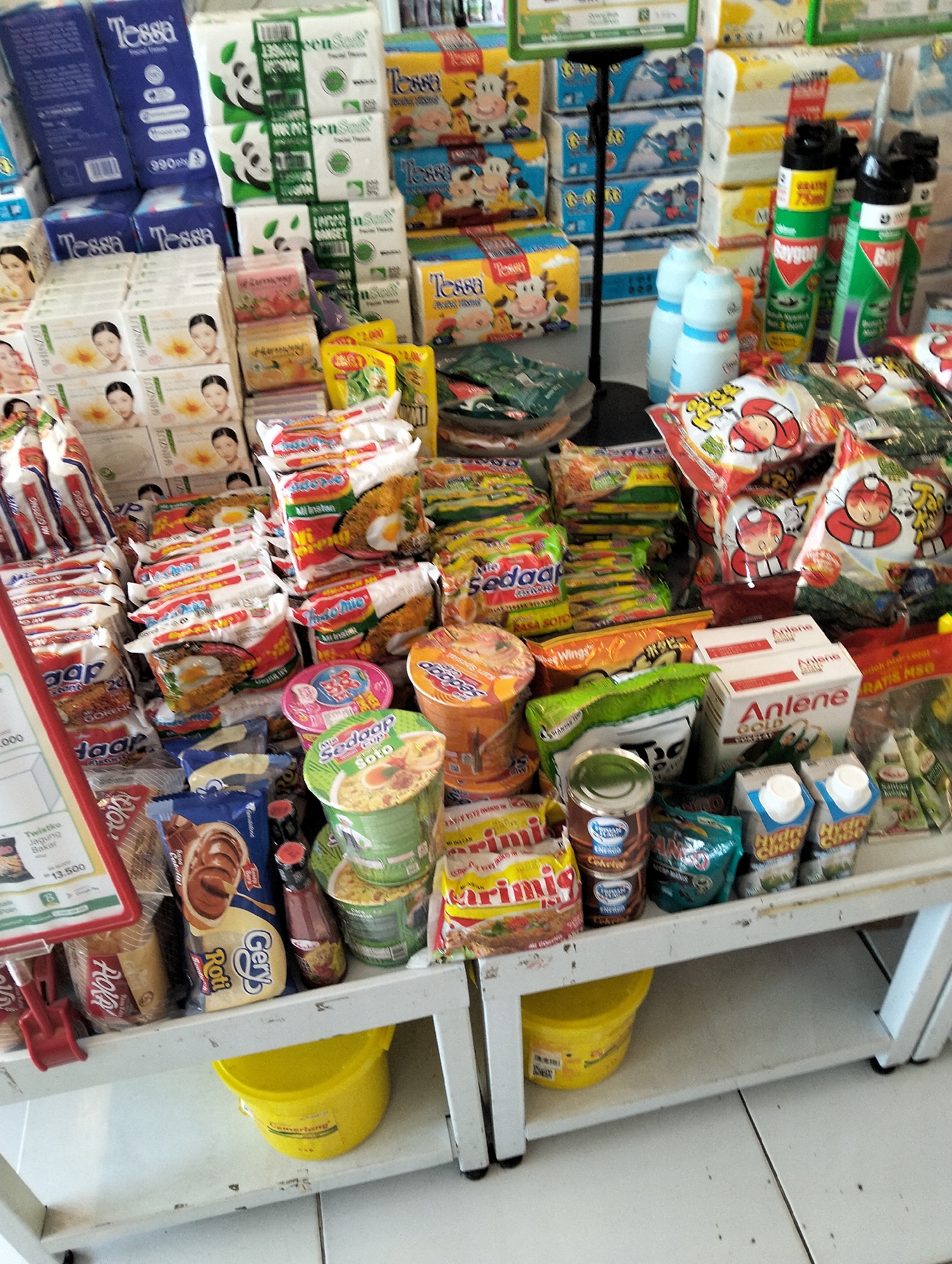 The image shows a retail shelf display in a store in Muncar, Indonesia. The shelves are filled with a variety of consumer goods, primarily food products and toiletries. Prominently displayed are several stacks of packaged instant noodles in cup and bag formats, such as Indomie and Sedaap brands, featuring colorful packaging with images of food. There are also packages of facial tissues, some with panda imagery, and boxes of soap or skincare products. In the background, aerosol cans, possibly insect repellent, are visible. Smaller items like small bottles of sauces or condiments, and cartons of milk or juice are also present. The shelves are made of white metal, and the overall environment appears to be a well-stocked convenience store or supermarket aisle. No people are visible in the image.