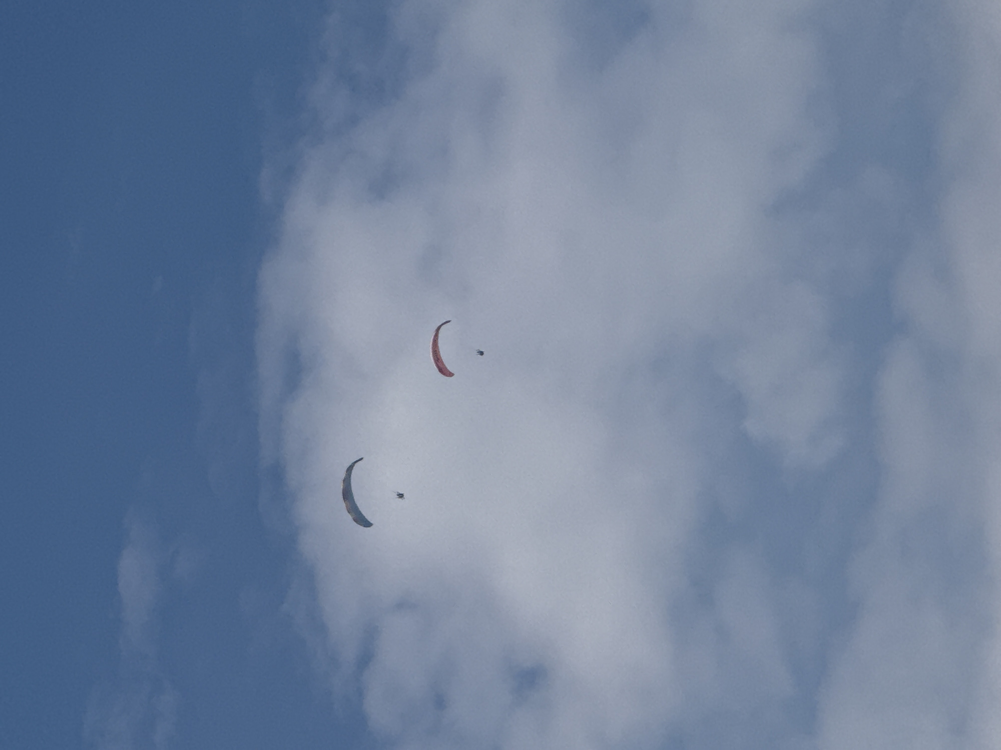 The image shows two paragliders in the sky above Bečići, Montenegro. The paragliders are at different altitudes and distances from the camera. The sky is blue with some clouds. The paraglider on the left is higher up and closer to the camera, while the paraglider on the right is lower and further away. The paragliders appear to be enjoying the scenery.