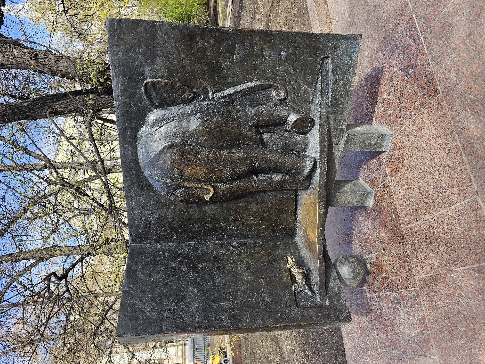The image depicts a bronze monument of Mikhail Bulgakov sitting at a desk, placed on a red granite base, possibly in Moscow, Russia. The statue shows Bulgakov in a seated position, seemingly writing, with a small dog figurine on the edge of his desk. The monument is positioned in an outdoor setting with trees and the sky visible in the background.
